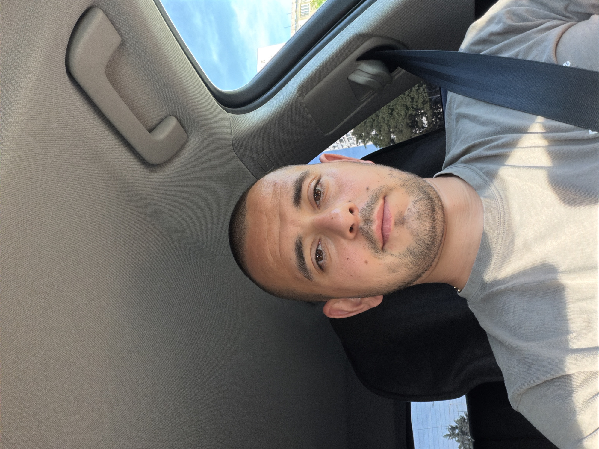 A man is seated in the driver's side of a car, facing forward and looking at the camera. He has a shaved head, a short beard, and is wearing a light gray t-shirt. A black seatbelt is fastened across his chest. The interior of the car is visible, including the roof lining, a grab handle, and a portion of the dashboard. Sunlight streams into the car, casting shadows on the man and the interior. Outside the car, trees and buildings are visible under a blue sky. The location is identified as being in Burgas, Bulgaria.