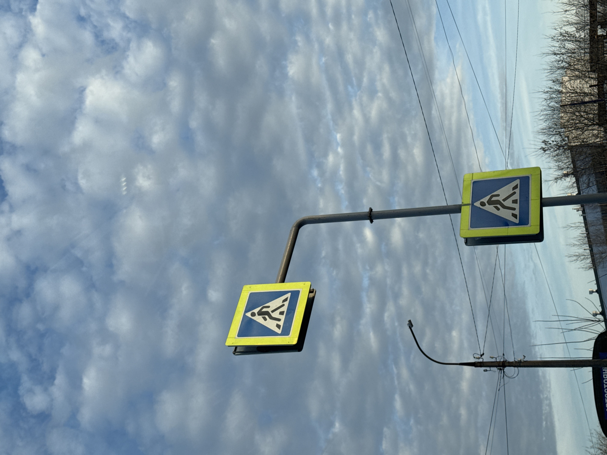 The image shows two road signs indicating a pedestrian crossing in Chaikovsky, Russia. The signs are mounted on a grey metal pole against a cloudy sky. The sign on the right is closer to the foreground and features a yellow border with a blue background depicting a walking figure within a triangle. The second sign is further away and has the same design. A street lamp is visible extending from a pole behind the road signs, and tree branches are visible in the background. Power lines run across the sky. The image is taken from a low angle.