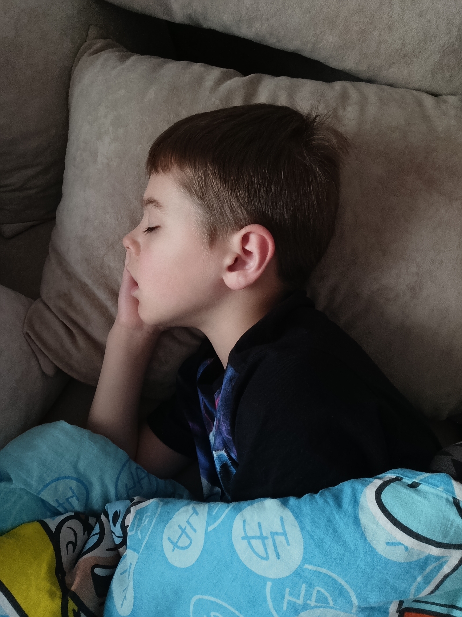 A young boy is sleeping on a beige upholstered couch in what appears to be a home setting. He is lying on his side, with his head resting on a pillow. His left hand is raised to his face. He is covered by a light blue blanket with white and yellow cartoon characters. He is wearing a black t-shirt. The location is Chaikovsky, Russia.