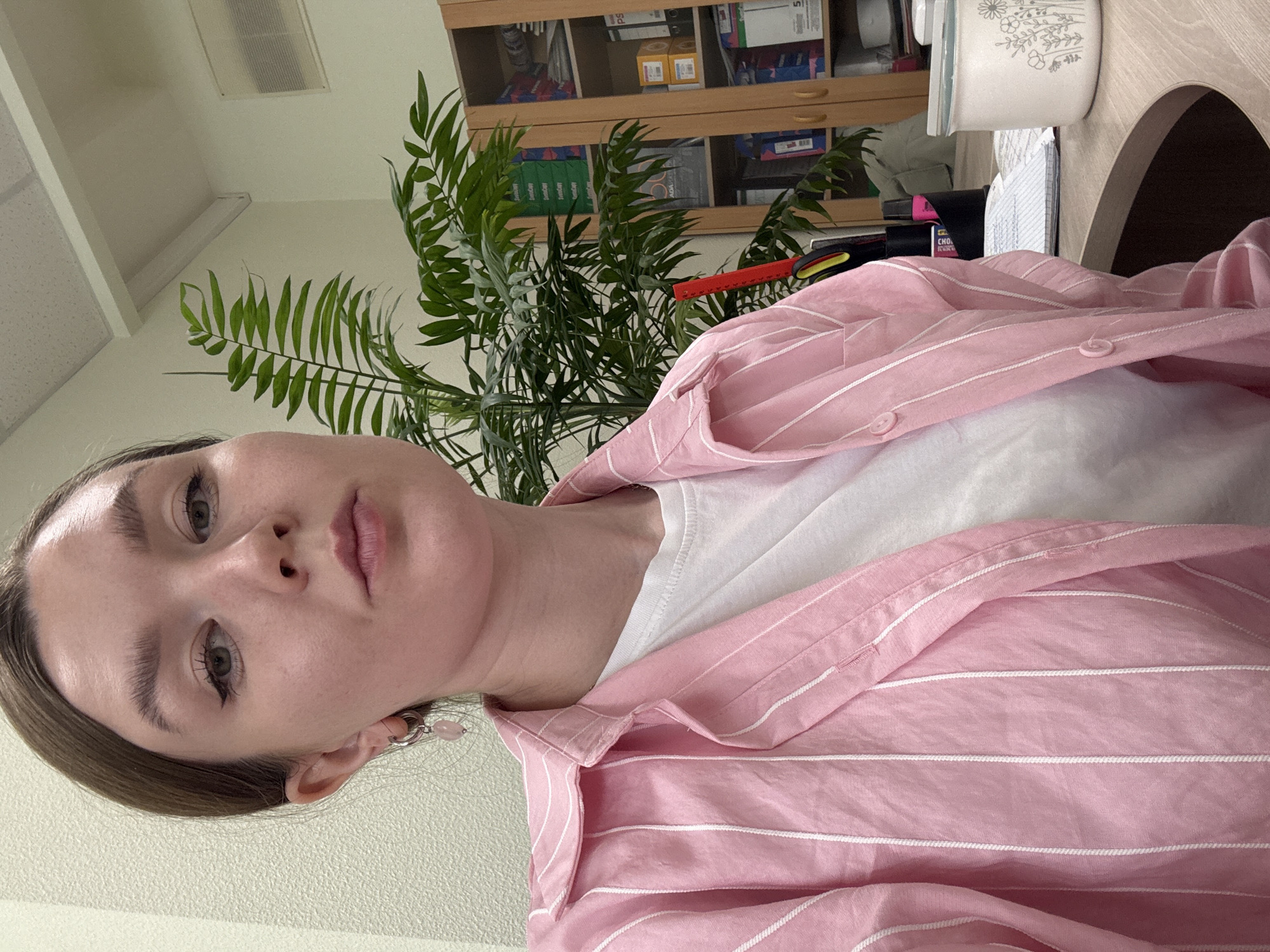 A selfie-style photograph, rotated 90 degrees counter-clockwise, captures a young person from a low-angle perspective. The individual, likely female, has light brown hair and is looking directly at the camera with a neutral expression. She is wearing a pink button-up shirt with thin white stripes over a white t-shirt. Silver hoop earrings with small pink beads are visible on her left ear.
The background suggests an indoor setting, likely an office or study room. A large green leafy plant, possibly a parlor palm, is prominent behind the person's right shoulder, extending towards the upper right and left of the frame. Behind the plant, a wooden shelving unit with several compartments holds various items, including binders, files, and boxes. Partially visible text on some boxes includes "eventCap", "ADA", and "PS". A red and black object, possibly a utility knife or lighter, rests on the shelves near the plant.
To the right, a white container with a delicate floral pattern is placed on a shelf. The ceiling features a rectangular vent and a fluorescent light fixture. The overall lighting is soft and even, suggesting indoor artificial light or indirect daylight.