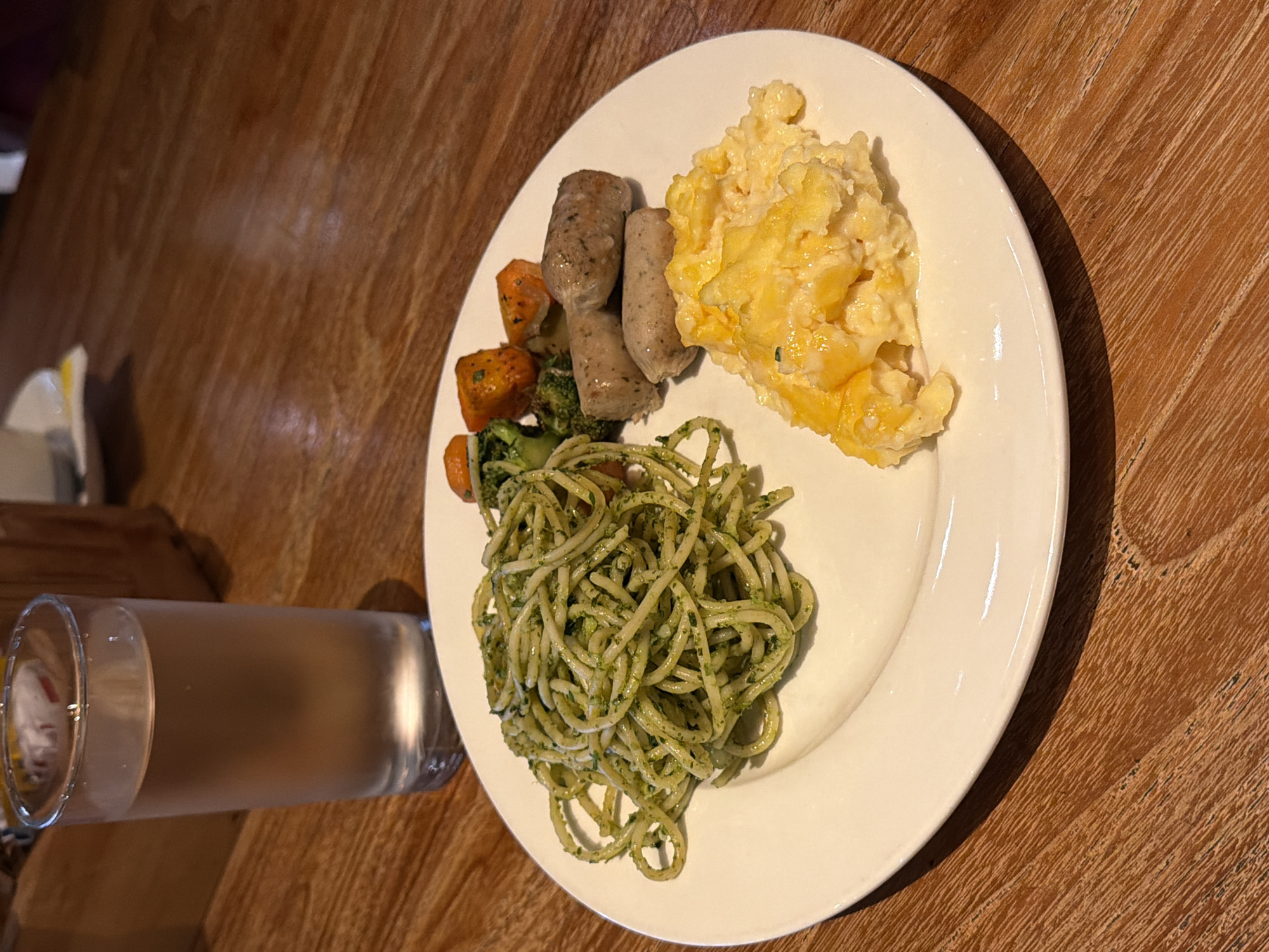 A white plate of food sits on a wooden table. The plate contains spaghetti with pesto, scrambled eggs, three sausages, and a mix of broccoli and roasted carrots. To the left of the plate is a tall glass of water. The scene is likely a restaurant in Kuta, Indonesia, given the context provided. There are no people visible in the image.