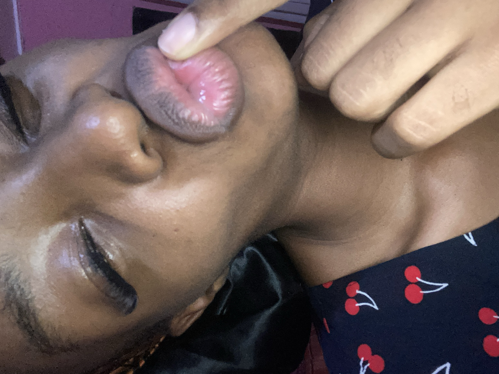 A close-up, angled portrait of an individual with dark brown skin, captured indoors in Jos, Nigeria. The subject's head is tilted, with their right eye partially closed or squinted, featuring prominent dark eyelashes. The nose is visible from the bridge down. Their lips are puckered into a "kissing" pose, with the inner surface of the lower lip appearing a lighter pink. The index finger of the subject's right hand is pressed against the center of the lower lip. A dark, shiny, possibly satin head covering is visible on the left side of the head, with hints of braided hair beneath. The subject is wearing a dark garment patterned with red cherries and white stems, visible on the lower left. The background is out of focus, primarily dark with a vertical strip of bright pink light on the right.
