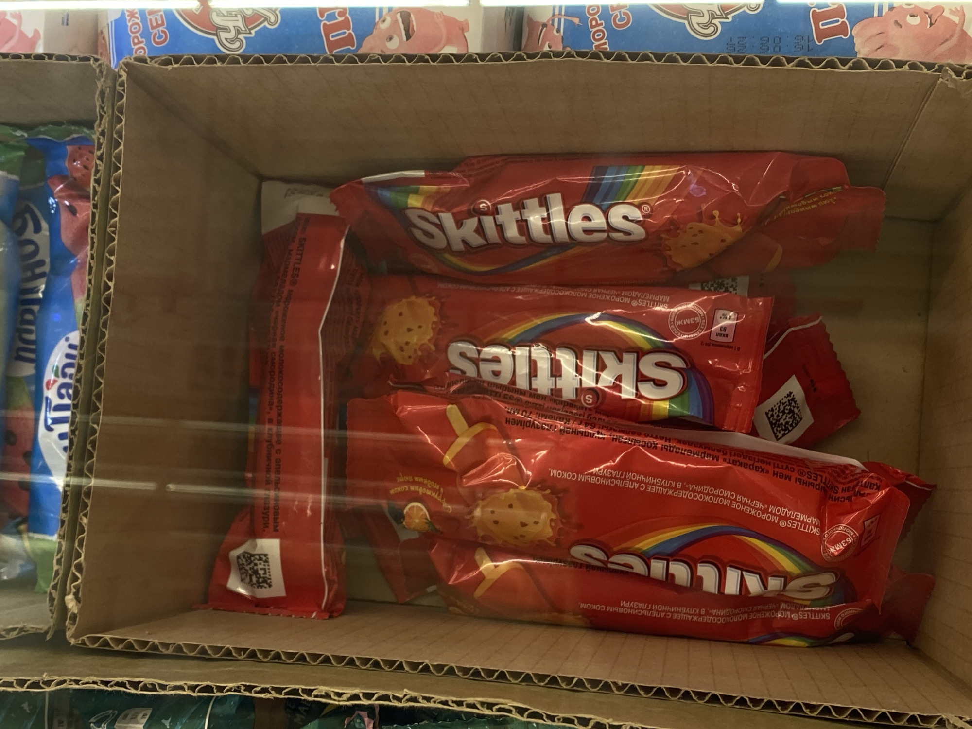 The image shows a cardboard box containing several red packages of Skittles candy. The packages have the Skittles logo and rainbow design. Some of the text on the packages is in Cyrillic. The box is placed in what appears to be a store or warehouse setting, with other products visible in the background. The lighting is artificial. The image is shot from a slightly elevated angle, looking down into the box.