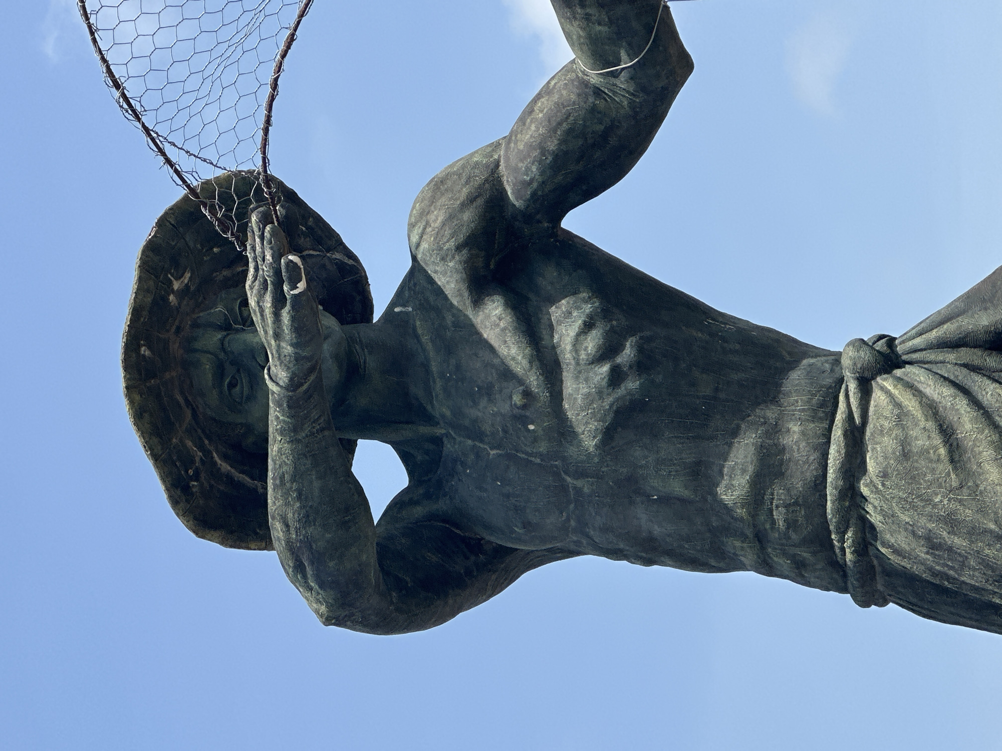 This image captures a bronze statue of a man from a low-angle perspective, set against a clear, bright blue sky with minimal, wispy clouds. The original image is rotated 90 degrees clockwise, making the statue appear horizontal, but it depicts a standing or striding figure.
The statue has a weathered, dark bronze finish with visible greenish patination, indicating age and outdoor exposure. The man is depicted with a muscular physique, unclothed from the waist up, showcasing detailed musculature in his arms, chest, and abdomen. A bronze cloth is draped around his lower body, forming a skirt-like garment secured at the waist.
He wears a wide-brimmed hat or head covering. Both of his hands are raised to hold a large, somewhat rectangular net made of wire mesh with hexagonal cells, attached to a thicker, curved frame. The net appears rustic and substantial. The man's face, partially visible beneath the hat and between his hands, shows a focused or solemn expression.
The bright, even lighting suggests it is daytime, likely mid-day or early afternoon, with strong sunlight. There is no visible text in the image. The statue is situated in Badagry, Nigeria.