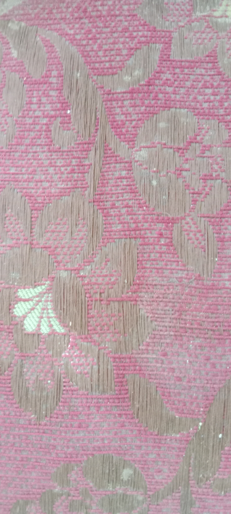 The image shows a close-up of a pink and beige patterned fabric, likely a textile or upholstery. The pattern consists of floral or leaf-like shapes in a lighter beige color against a pink grid-like background. The texture of the fabric is visible, with fine threads forming the pattern. There are also small specks of white or light-colored dust or debris scattered across the surface. The lighting appears to be natural, casting subtle shadows and highlighting the texture.
This image does not contain any information that would allow for its localization within the city of Tuban, Indonesia, or any other specific location. It is a generic close-up of a patterned textile.