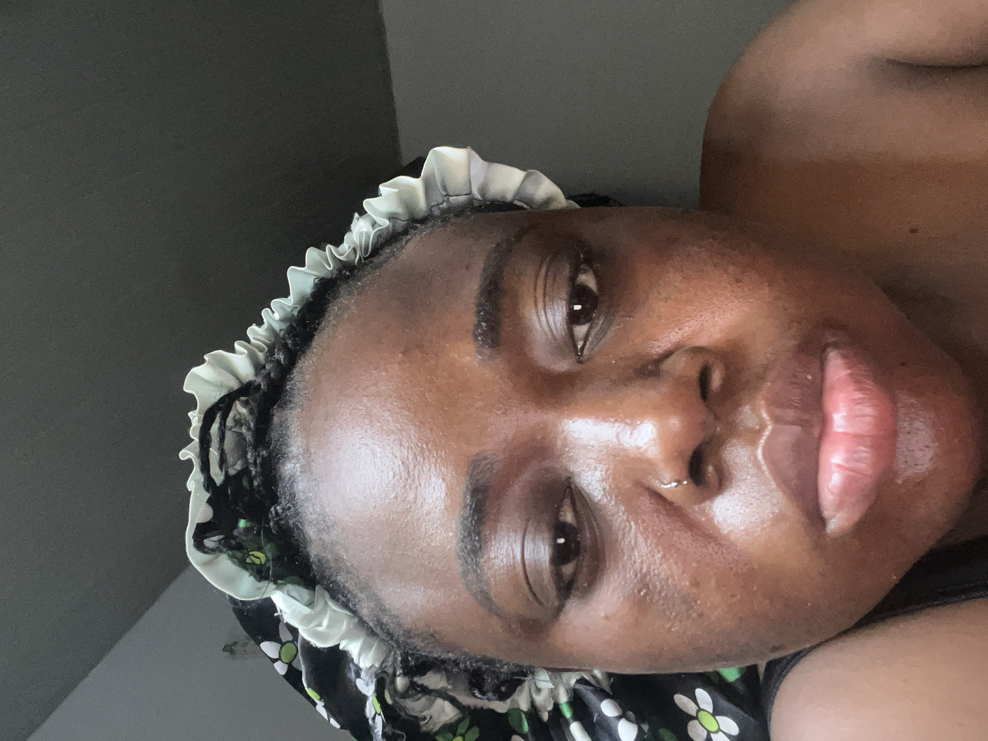 A close-up photograph depicts a Black woman with dark hair partially covered by a floral-patterned bonnet with a white ruffled edge. She has clear skin, full lips, a nose ring, and dark eyebrows. The background is a muted gray. The location is unknown and cannot be determined from the image.