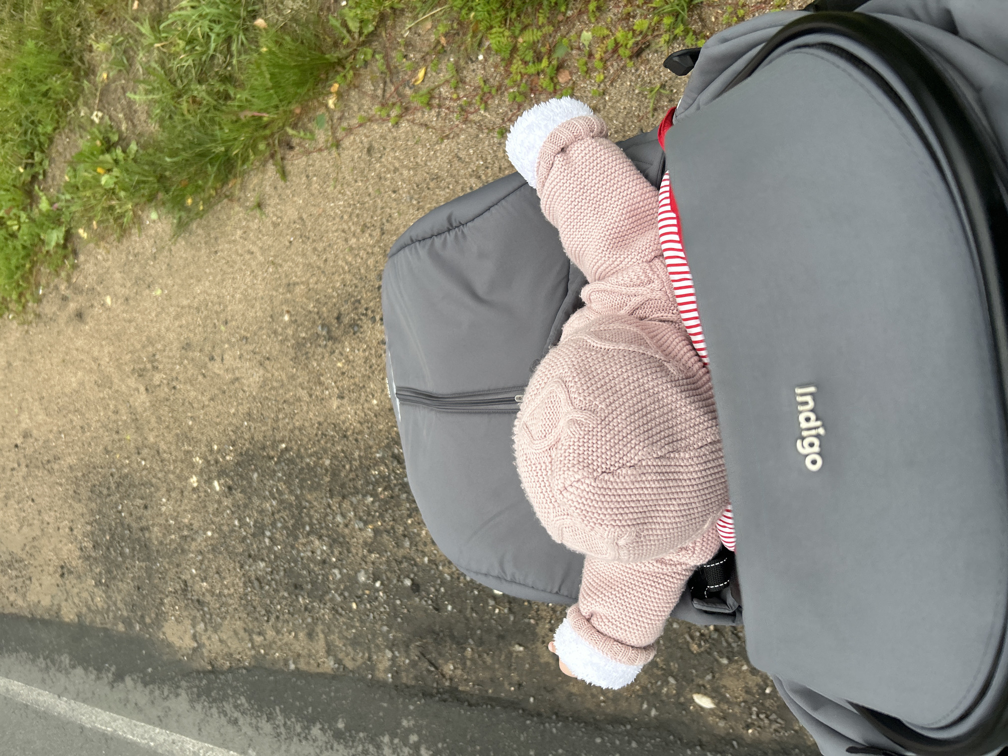 A first-person perspective shot from a stroller shows a baby wearing a pink knitted hat and sweater, with white trim on the sleeves, being pushed along a path. The path appears to be made of gravel and dirt, with some green grass growing along the side. To the left of the path, there is a grey asphalt surface with a white line marking its edge. The stroller itself is grey, and the brand name "Indigo" is visible on the front. The background is blurred, suggesting movement, and the lighting indicates it is daytime. The location is a paved or semi-paved path in a natural outdoor setting, likely a park or residential area.