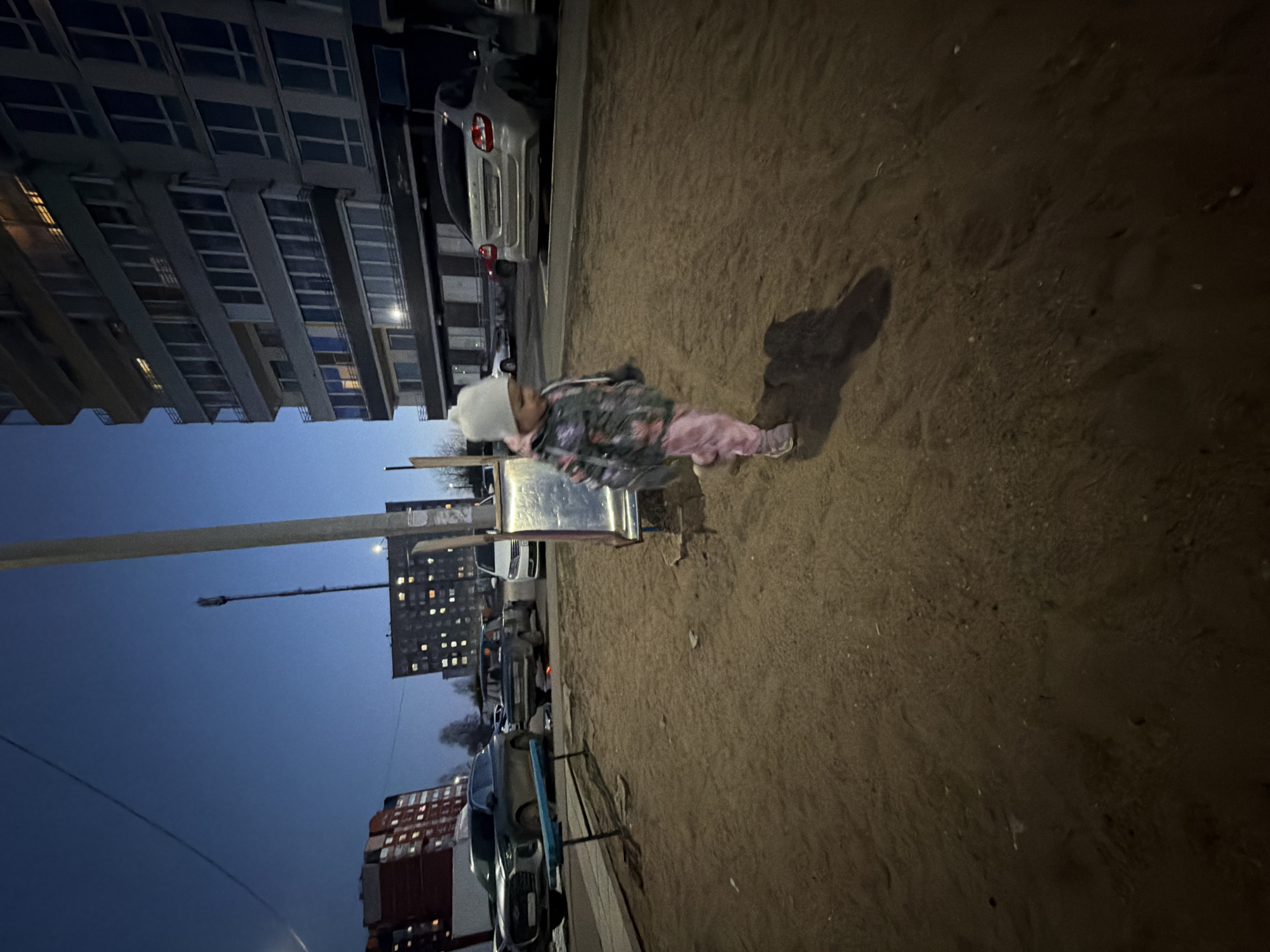 A child in a grey patterned jacket, pink pants, and a white hat stands in a sandbox in Chaikovsky, Russia. The sandbox is located near a tall apartment building. A street light pole is visible in the background. Several parked cars and other buildings are nearby. The time of day appears to be evening or early morning.