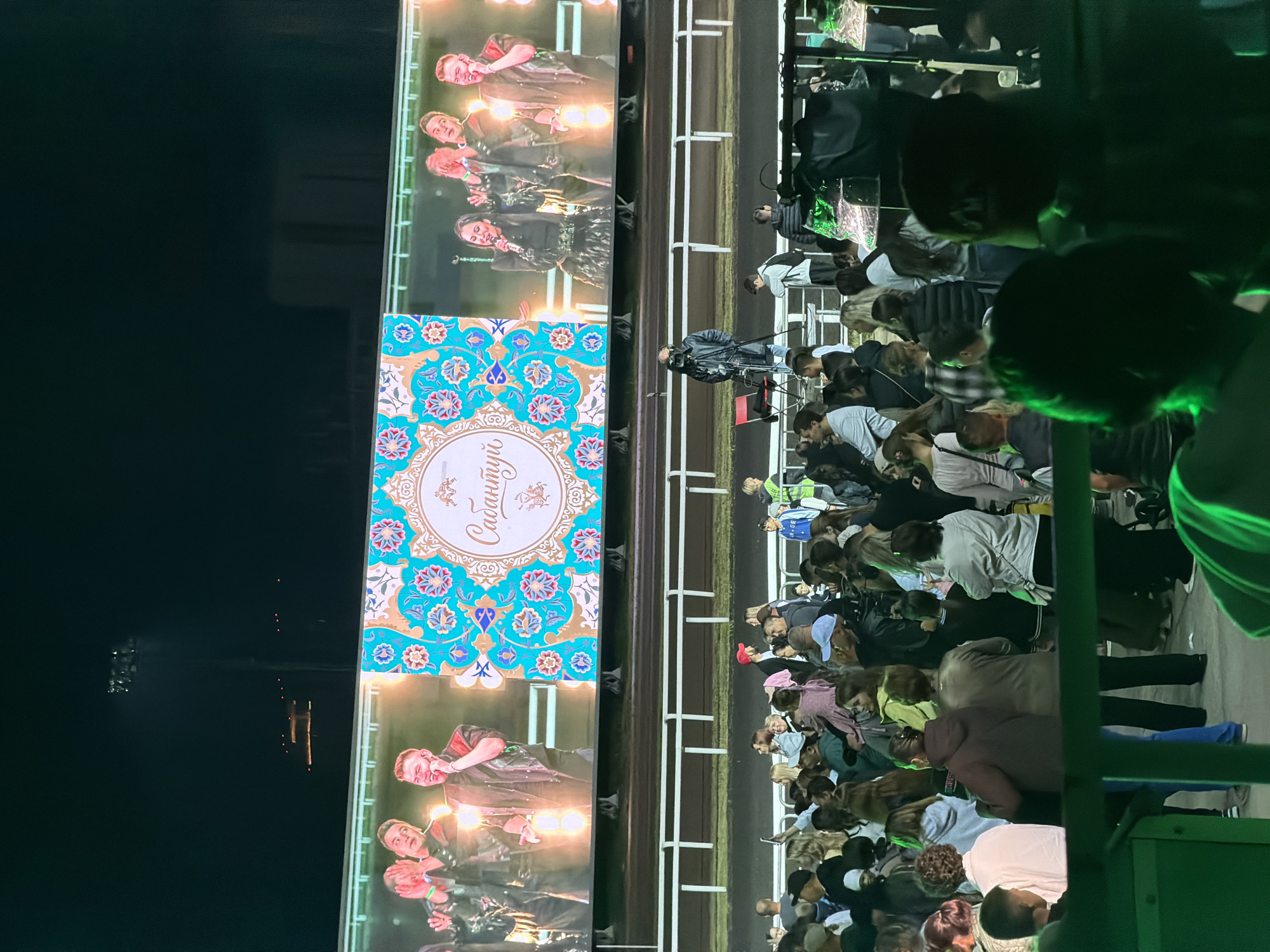 In an outdoor, nighttime stadium setting, a large crowd of people watches a massive LED screen displaying a live performance. The screen shows a group of singers on stage, with the center featuring a decorative graphic with text that reads "Сабантиу" in ornate script. The crowd is gathered in tiered seating areas and standing in a viewing area behind a fence, with some individuals holding up cell phones to record the event. In the foreground, the backs of heads of audience members are visible, obscured by dark green fencing. A camera operator with a professional video camera on a tripod is positioned on a raised platform in front of the screen, filming the performance. The environment appears to be a sports stadium or concert venue, with stadium lighting visible in the dark background.
