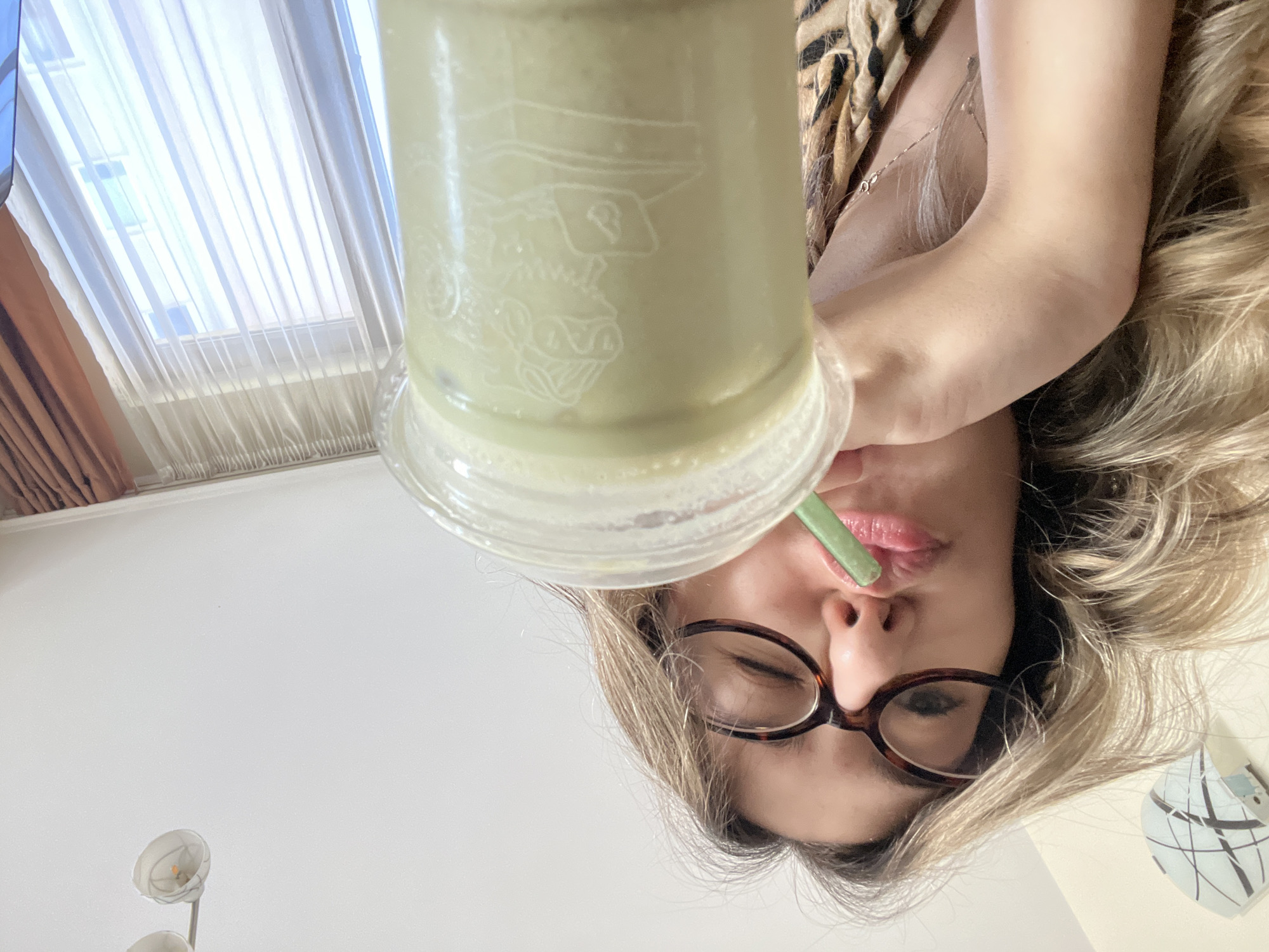 The image is a rotated perspective shot of a woman drinking a light green smoothie. She is wearing brown-framed glasses and has blonde hair. She is wearing a cheetah print top. The smoothie is in a clear plastic cup with a green straw. A drawing of a man with a hat on the glass. The background includes a window with sheer curtains and a floral lamp. The exact location in Sofia, Bulgaria cannot be determined from this information.