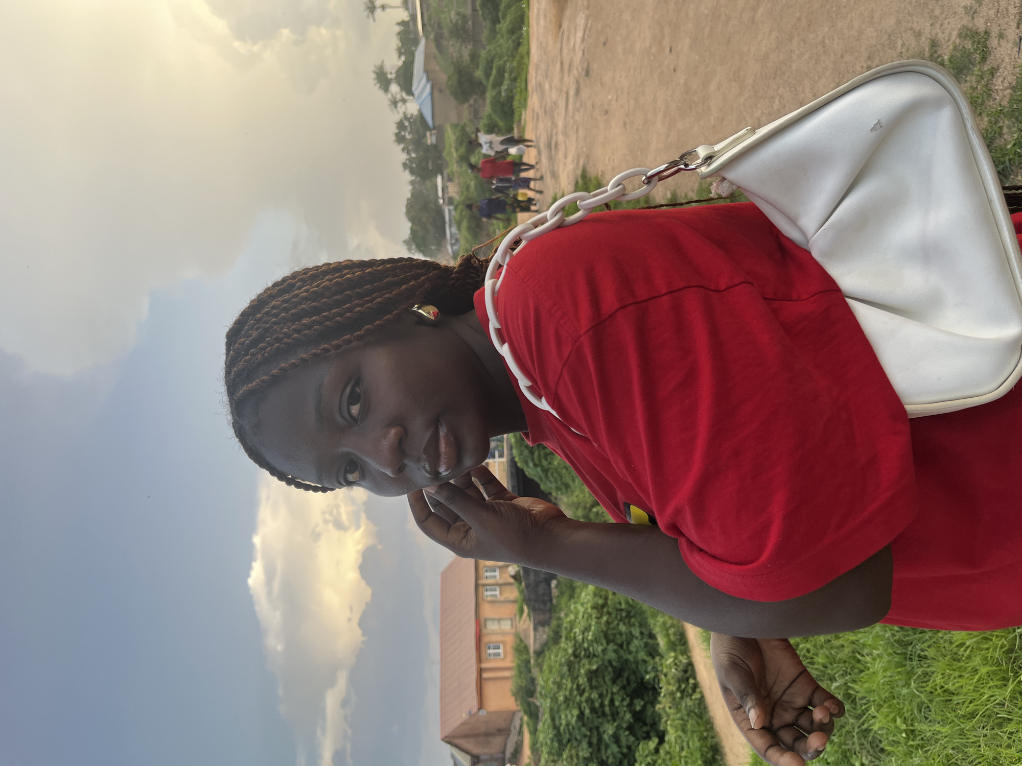 The media file shows a young Black woman posing outdoors. She has dark braided hair and is wearing a red t-shirt. A white purse with a white chain strap is slung over her right shoulder. Her left hand is raised to her chin, and she is looking towards the camera with a neutral expression.
The background suggests an outdoor setting in a rural or semi-rural area. There is a dirt path and green grass, with some buildings and trees visible in the distance. A group of people can be seen further in the background. The sky is cloudy with patches of sunlight, indicating it might be late afternoon or early morning. The overall lighting suggests a bright, possibly warm, day.
There is no visible text in the media file. The scene appears to be a casual portrait taken in Jos, Nigeria. The focus is on the woman, but the background provides context of her surroundings.
