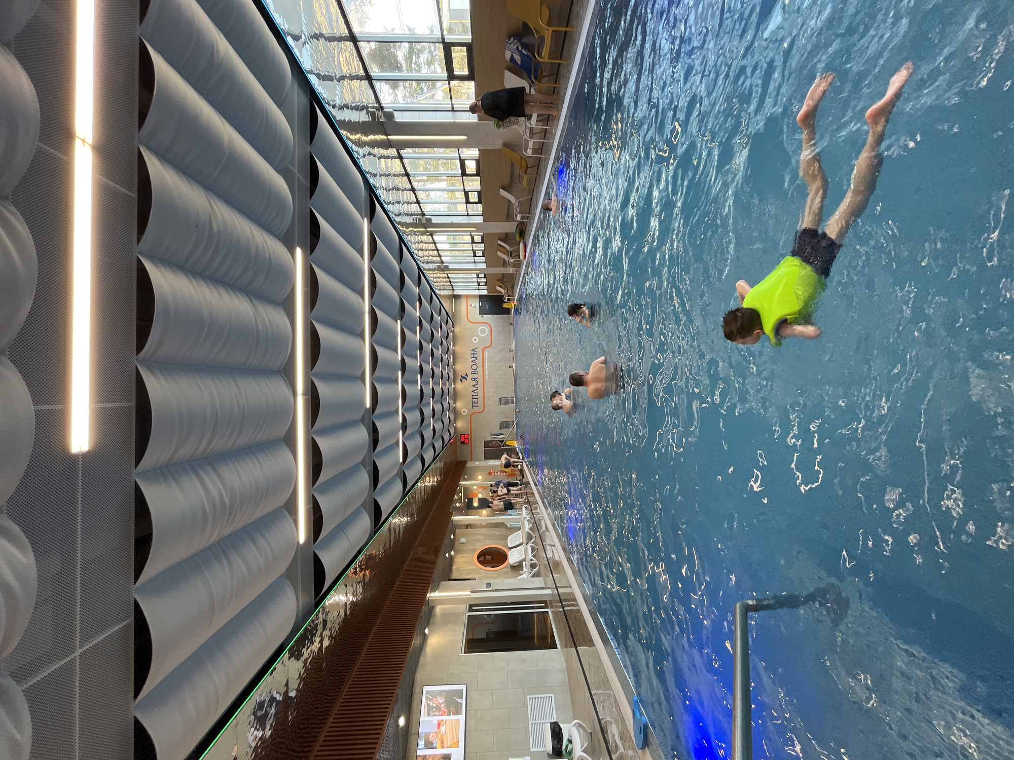 The image depicts an indoor swimming pool. A child wearing a neon green life vest and dark swim trunks is submerged in the water, appearing to be swimming or diving. Other people are present in the pool, including at least two adults who are swimming. Along the side of the pool, there are chairs and some people standing or sitting. The pool is located in Votkinsk, Russia, as evidenced by the signage "ТЕПЛАЯ ВОЛНА" (Teplaya Volna - Warm Wave). The pool area features a wall with a textured design of rounded shapes. There is a glass ceiling with natural light coming through. Artificial lights are also visible on the wall. The water in the pool is a clear blue color.