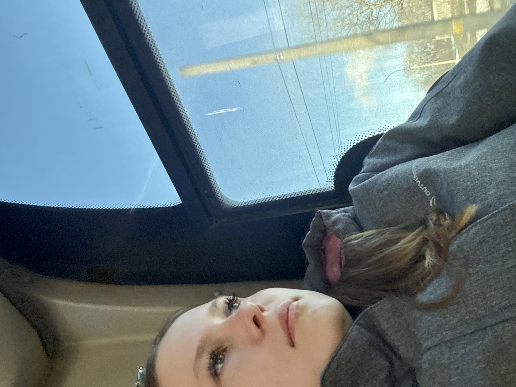 The image shows a person with fair skin, brown hair, and gray eyes, lying down inside a vehicle with a sunroof. The sunroof offers a view of a light blue sky with a few visible structures, including a yellow horizontal bar and trees, in Berezniki, Russia. The person is wearing a gray hooded sweatshirt with pink lining visible. The scene is brightly lit by daylight.