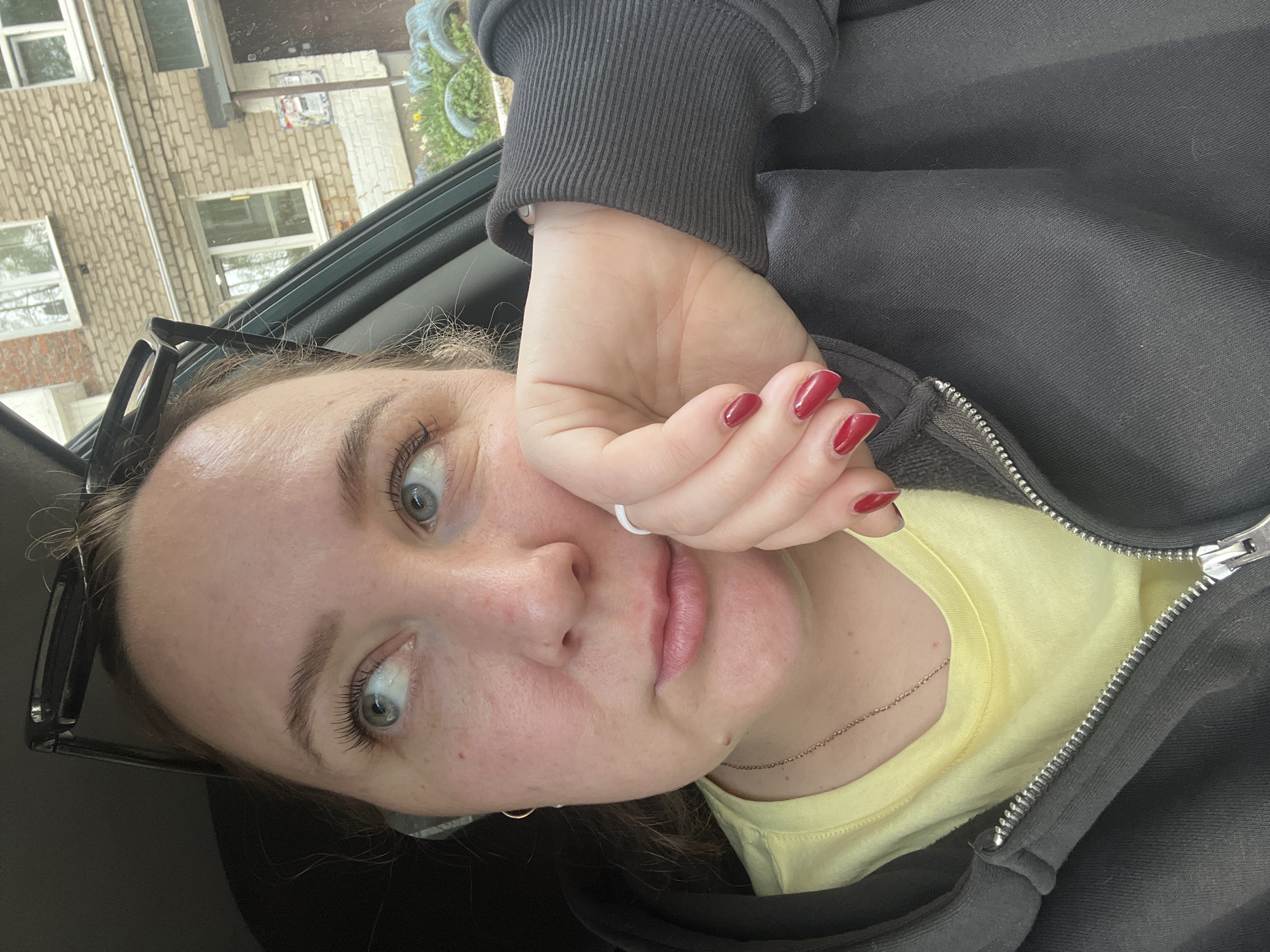 A pale-skinned woman with red nail polish is pictured from a close, low-angle point of view, seemingly inside a car. She is wearing a yellow shirt and a dark gray zip-up hoodie. Her left hand is near her face, with a white ring on one finger. A pair of eyeglasses is positioned next to her face. Outside the car window, a multi-story brick building is visible. According to metadata, the photo was taken in the city of Chaikovsky, Russia.