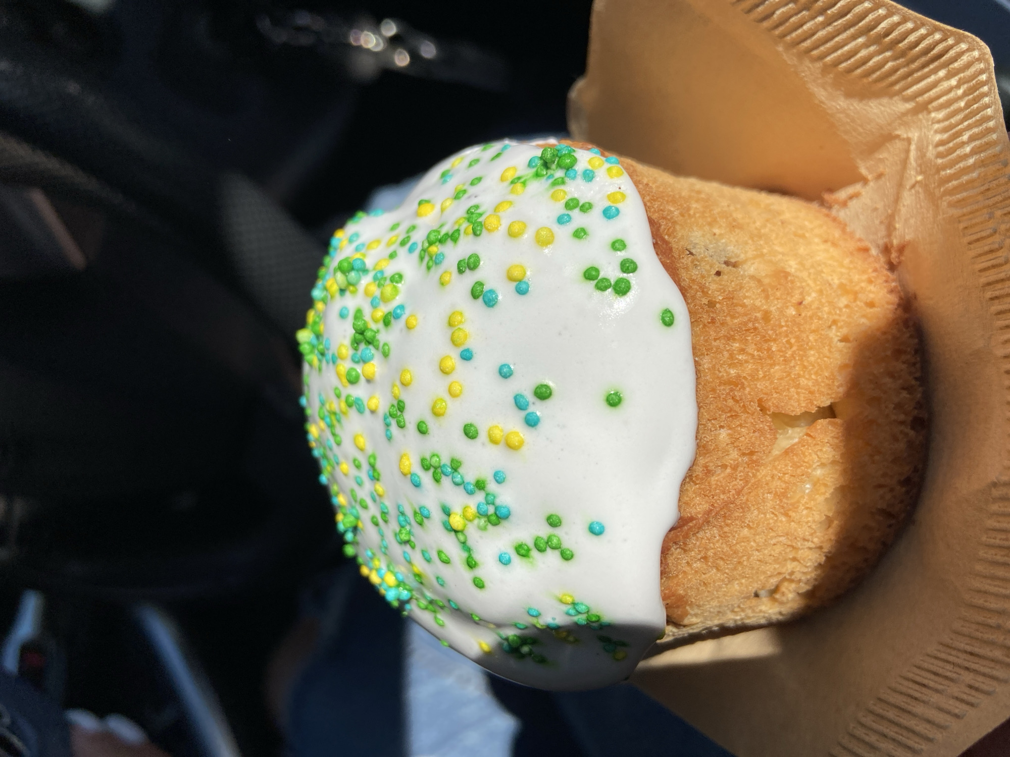 A pastry is half-covered in white icing and sprinkles of green, yellow, and blue. The pastry sits in a brown corrugated paper cup. The background contains dark surfaces and the edge of a blue fabric. The location of the image is unspecified.
