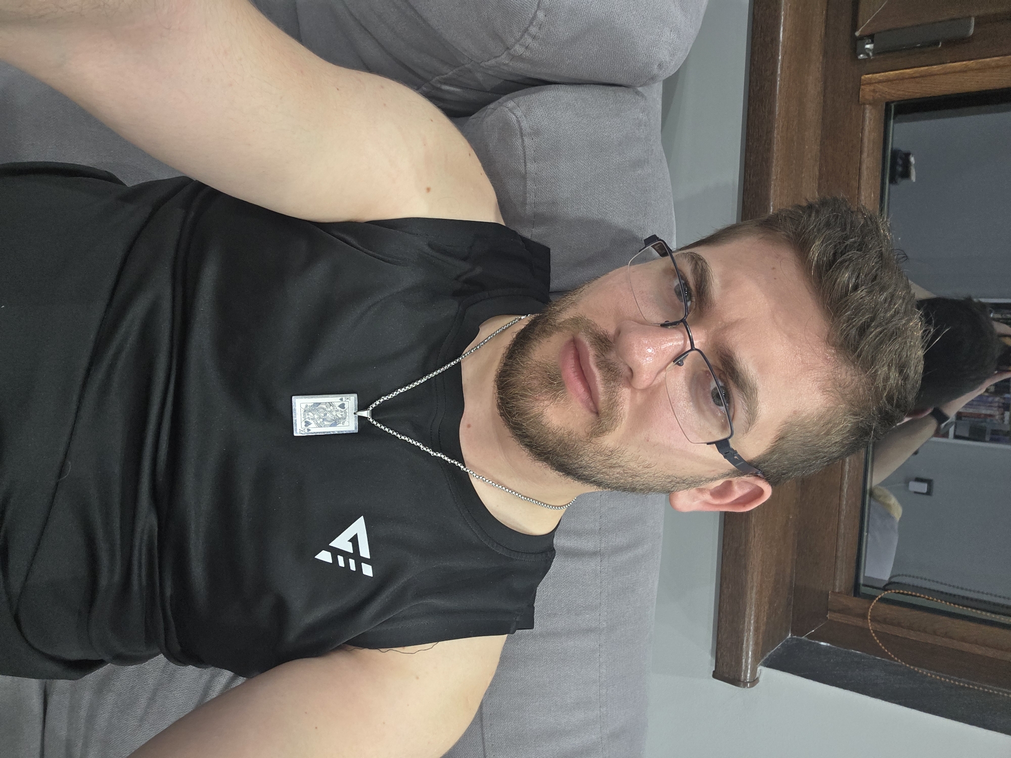 A man with a beard and glasses is lying on a gray couch. He is wearing a black tank top with a white logo on the left side and a silver chain with a pendant of a playing card, Queen of Spades. The background includes a window frame. It is unlikely I can determine the location as Yagodovo, Bulgaria, based solely on the image.