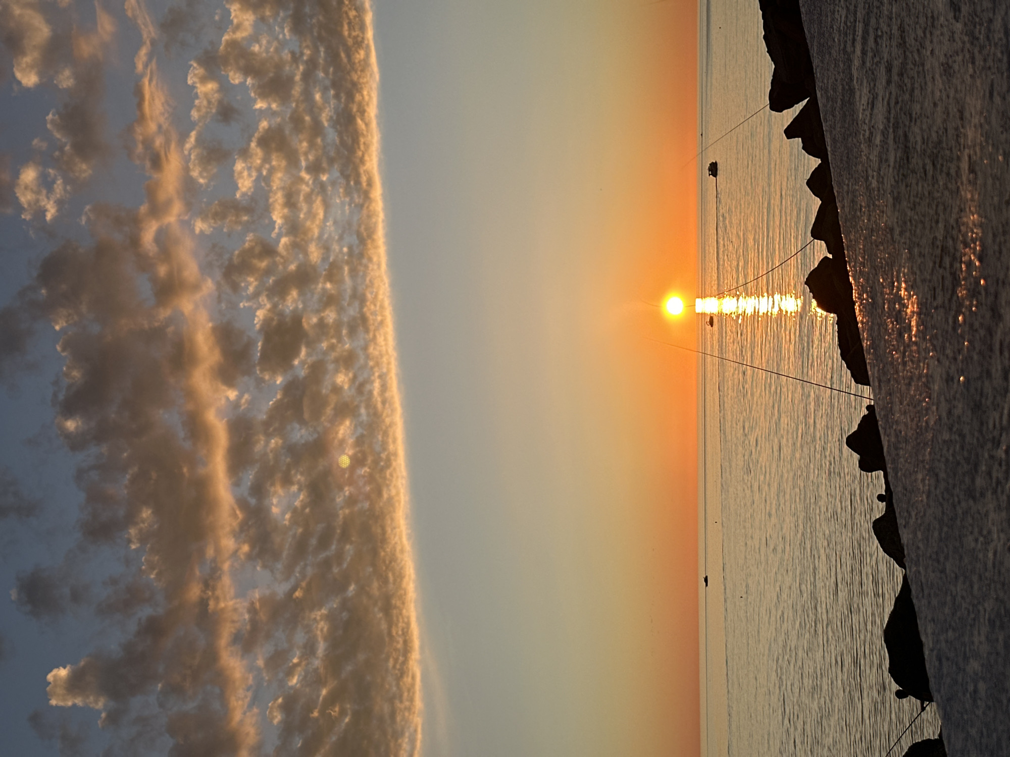The image depicts a sunset over the sea, with a rocky shoreline visible on the right side. A band of textured clouds occupies the left side of the frame. The sun is low on the horizon, its reflection stretching across the water. Several small, dark objects (possibly boats or fishing gear) are scattered on the water's surface. The location could be Sirius, Russia, given the coastal landscape and sunset orientation.