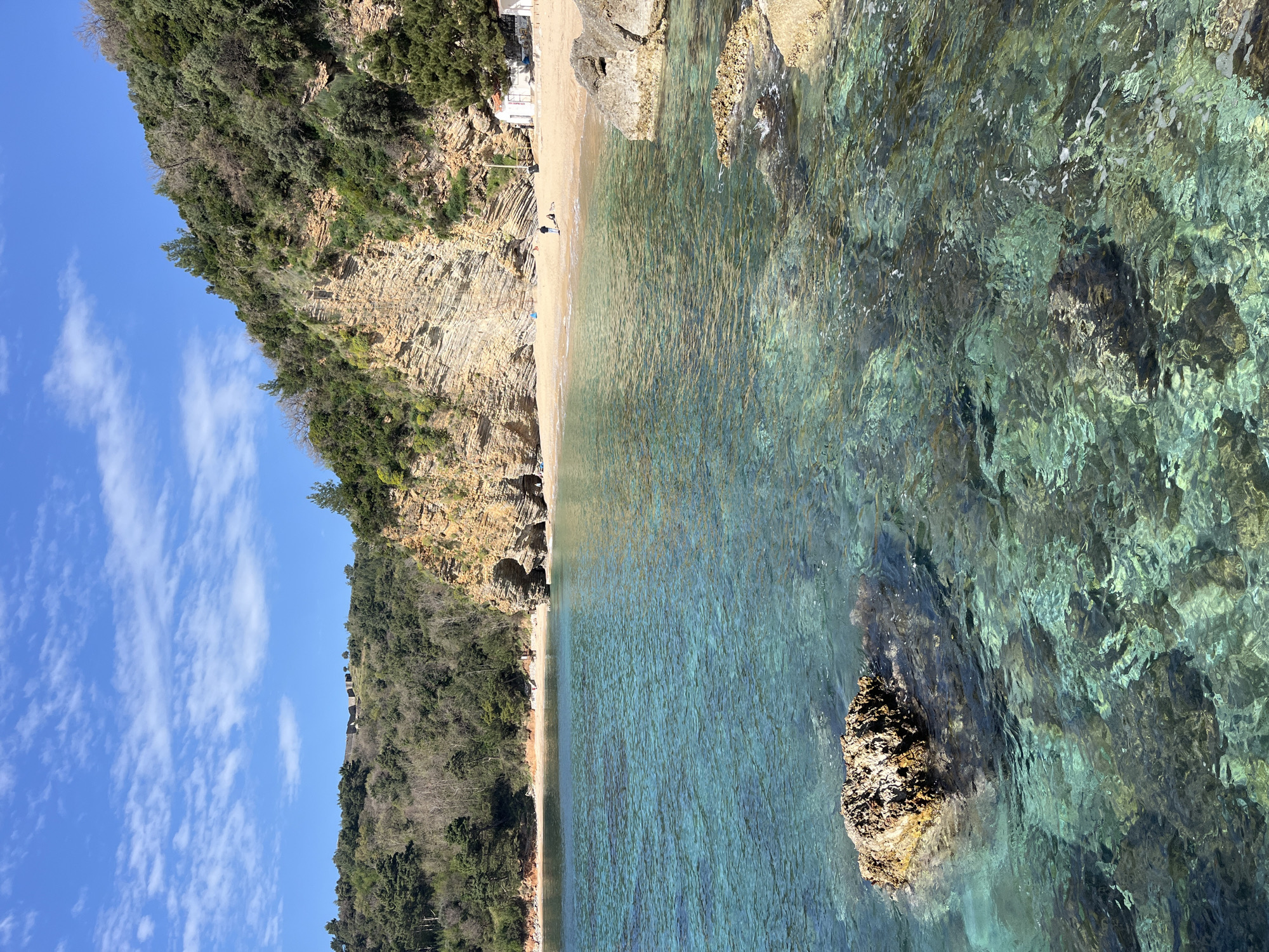 The image shows a beach near Budva, Montenegro, on a sunny day with a blue sky and light clouds. The beach is bounded by a rocky cliff covered in trees. The water is clear and turquoise in color, with visible rocks under the surface. There are a few people on the beach.