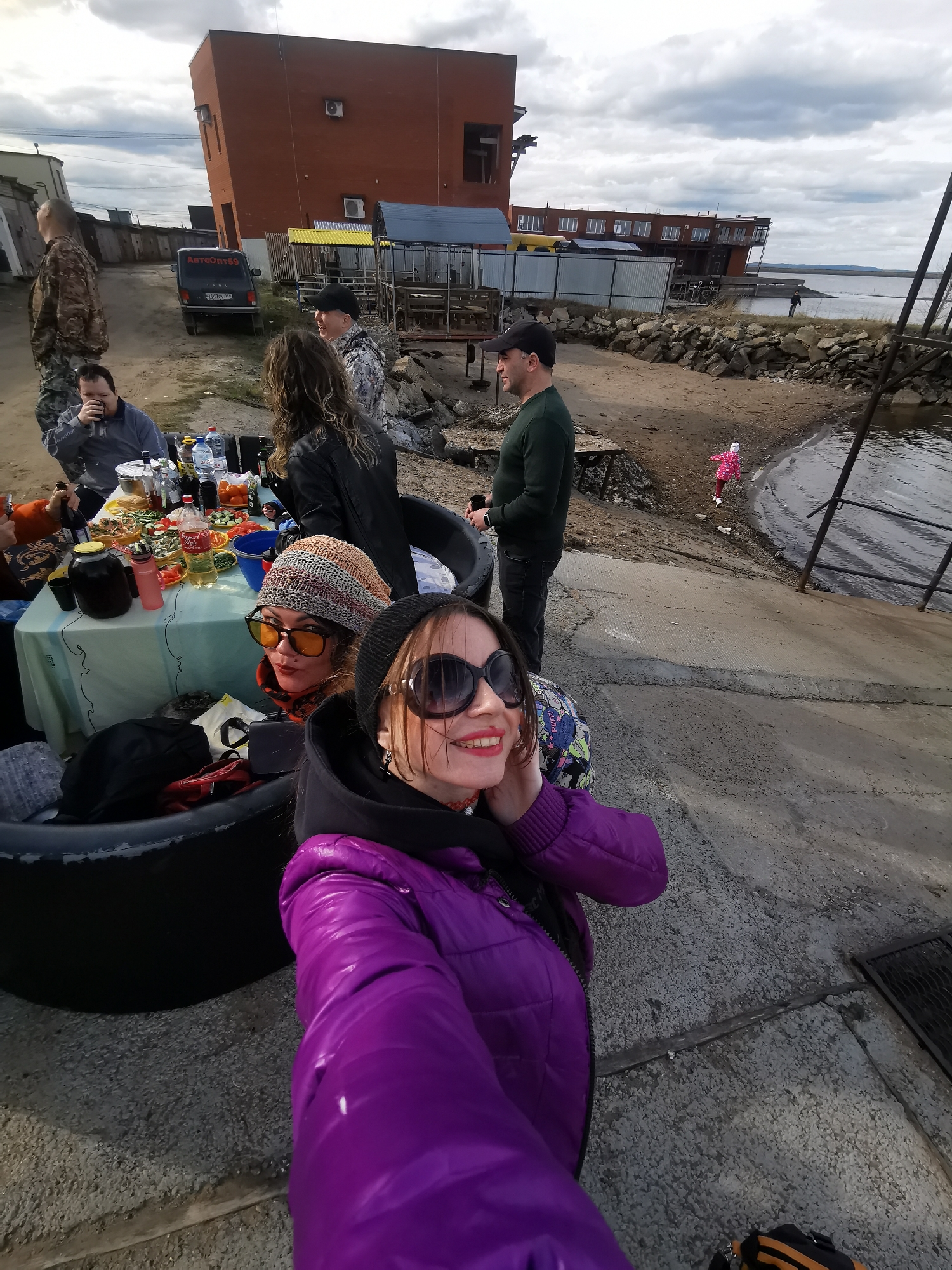 An outdoor gathering is depicted with several individuals enjoying a meal and socializing by a body of water under an overcast sky. In the foreground, two women are posing for a photo. The woman on the right wears a purple puffer jacket, a black beanie, and large sunglasses, smiling brightly with red lipstick. The woman to her left has a striped beanie, orange-tinted sunglasses, and an orange scarf, looking towards the camera with a slight smile. They are seated, possibly on large black tires.
Behind them, a table covered with a light blue tablecloth is laden with food and drinks. Visible items include various dishes with salads, bottles of Coca-Cola and Sprite, water bottles, a large dark jar, and a red thermos. Several adults are gathered around the table; one man in a grey top is seen drinking from a cup, and another man in a camouflage jacket is also present. Further back, a man in a dark green sweater and black cap stands near the water's edge, while another man in camouflage is visible on the left.
The setting appears to be a mix of concrete and dirt ground leading to a rocky shoreline and a large body of water, possibly a lake or sea. In the distance, a small child in a pink jacket is seen playing near the water. The background features several industrial-style buildings, including a distinctive multi-story brick building with sloped walls, and other smaller structures. A dark minivan with "АвтоОпт59" written on its rear window is parked on a dirt patch. A metal structure, possibly a pier or ramp, extends towards the water on the right. The weather is cloudy but with adequate daylight, suggesting either late morning or afternoon. The overall mood is casual and social.