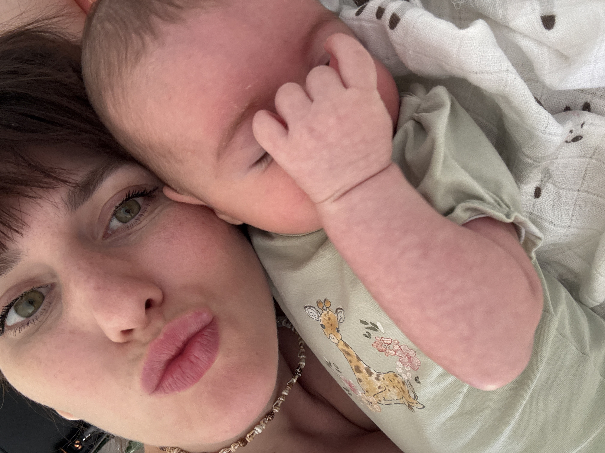 The media file is a close-up, high-angle selfie taken in a home setting. The primary subjects are a woman and a baby. The woman, positioned in the foreground on the left, has dark hair, green eyes, and is making a kissing gesture with her lips. She is wearing a necklace with a natural, beaded design. The baby, on the right, is nestled against the woman, with its head resting on her shoulder. The baby's skin is pink and it is wearing a light green onesie with a giraffe and floral print. A light, patterned blanket is visible in the background. The lighting appears to be natural and soft, suggesting daytime. The overall mood is intimate and affectionate, capturing a moment between mother and child. No visible text is present in the image.