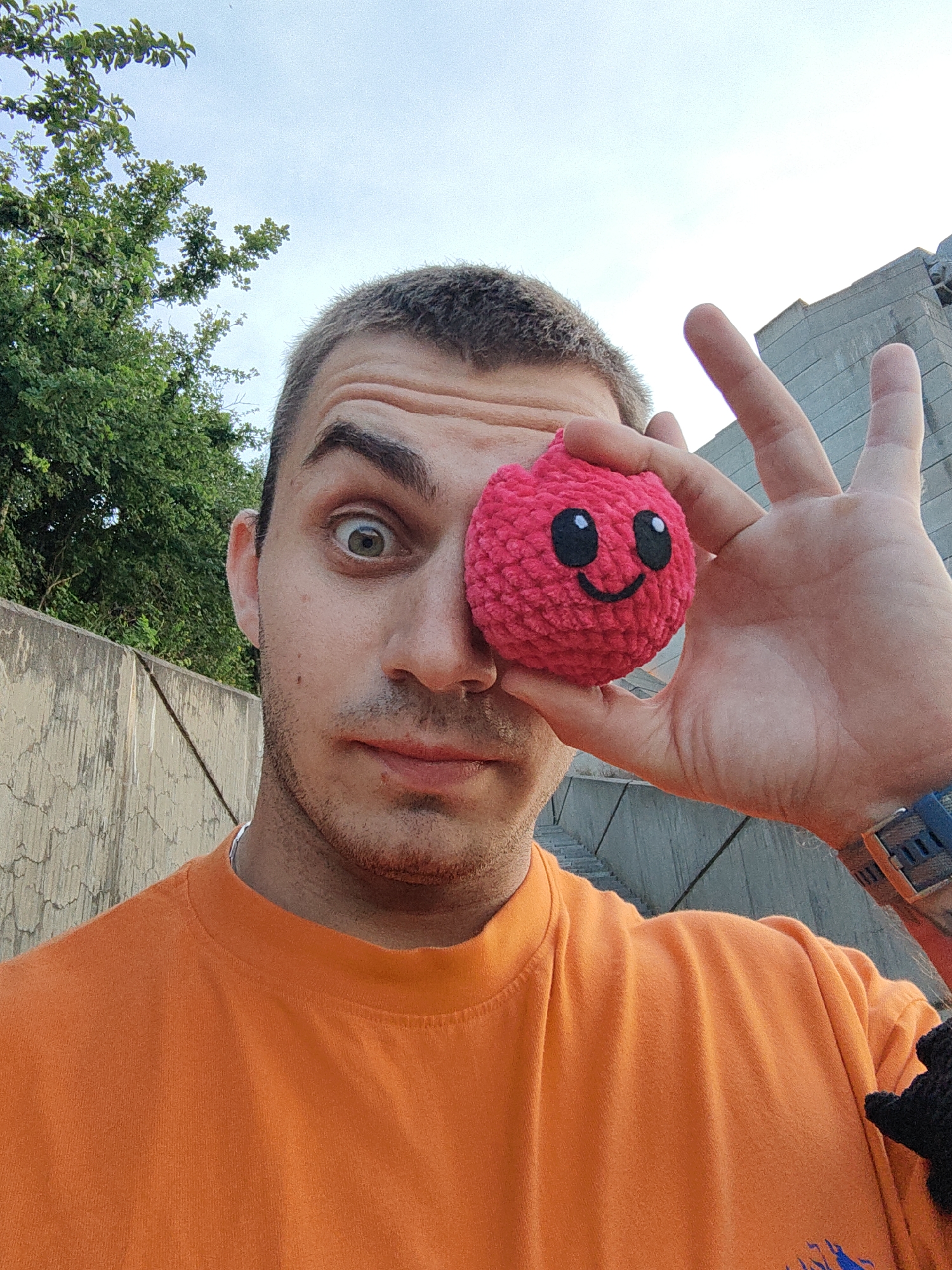 A young man with short, dark hair and a beard is taking a selfie outdoors, holding a small, pink, crocheted plush toy with embroidered eyes and a smiling mouth in front of his right eye. He is wearing an orange t-shirt. The background shows a concrete wall on the left and a modern building on the right, with trees and a pale blue sky above. The man's left eye is wide open, and he is looking at the camera. His right hand is raised, with two fingers extended in a peace sign, partially obscuring the plush toy. The image appears to have been taken at eye level, suggesting the man is holding the camera at arm's length.