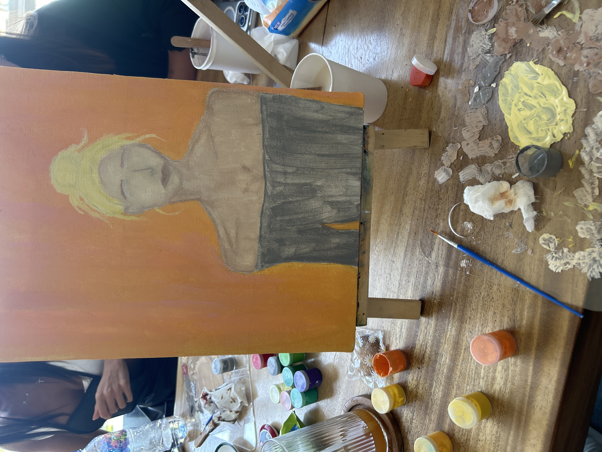 The image shows a partially completed painting on a canvas held by an easel. The painting is a portrait of a person with light hair and a gray garment, set against an orange background.
Scattered around the easel and canvas are various painting supplies: small containers of paint in different colors (yellow, orange, red, green, blue, white, brown), paint brushes, a palette with mixed paint colors, and cups (presumably for water or cleaning brushes). There also appears to be some tissues or cloth fragments used for cleaning.
Parts of two individuals are visible: one behind the painting near the top of the image, and another at the bottom-left corner of the image.
The environment is a wooden table, suggesting an indoor location for painting. The location within Kuta Selatan, Indonesia is not discernible from the image.