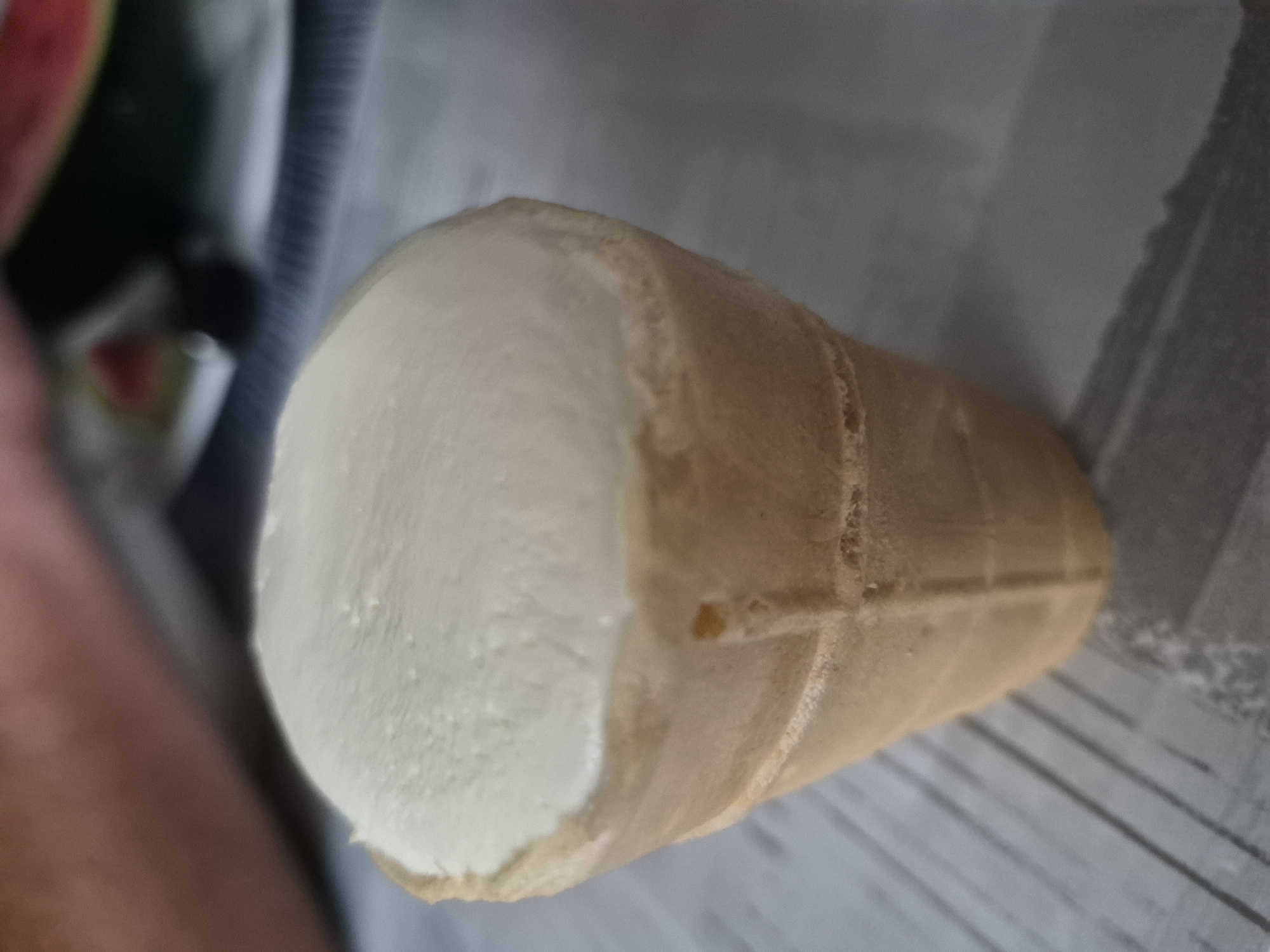 A close-up shot shows a single vanilla ice cream cone in an ice cream cone holder. The ice cream is a light, creamy white and appears to be soft-serve. The cone is a light golden brown and has a ribbed texture. The background is blurred and consists of a gray striped surface and hints of what appears to be watermelon. The image is likely taken indoors.