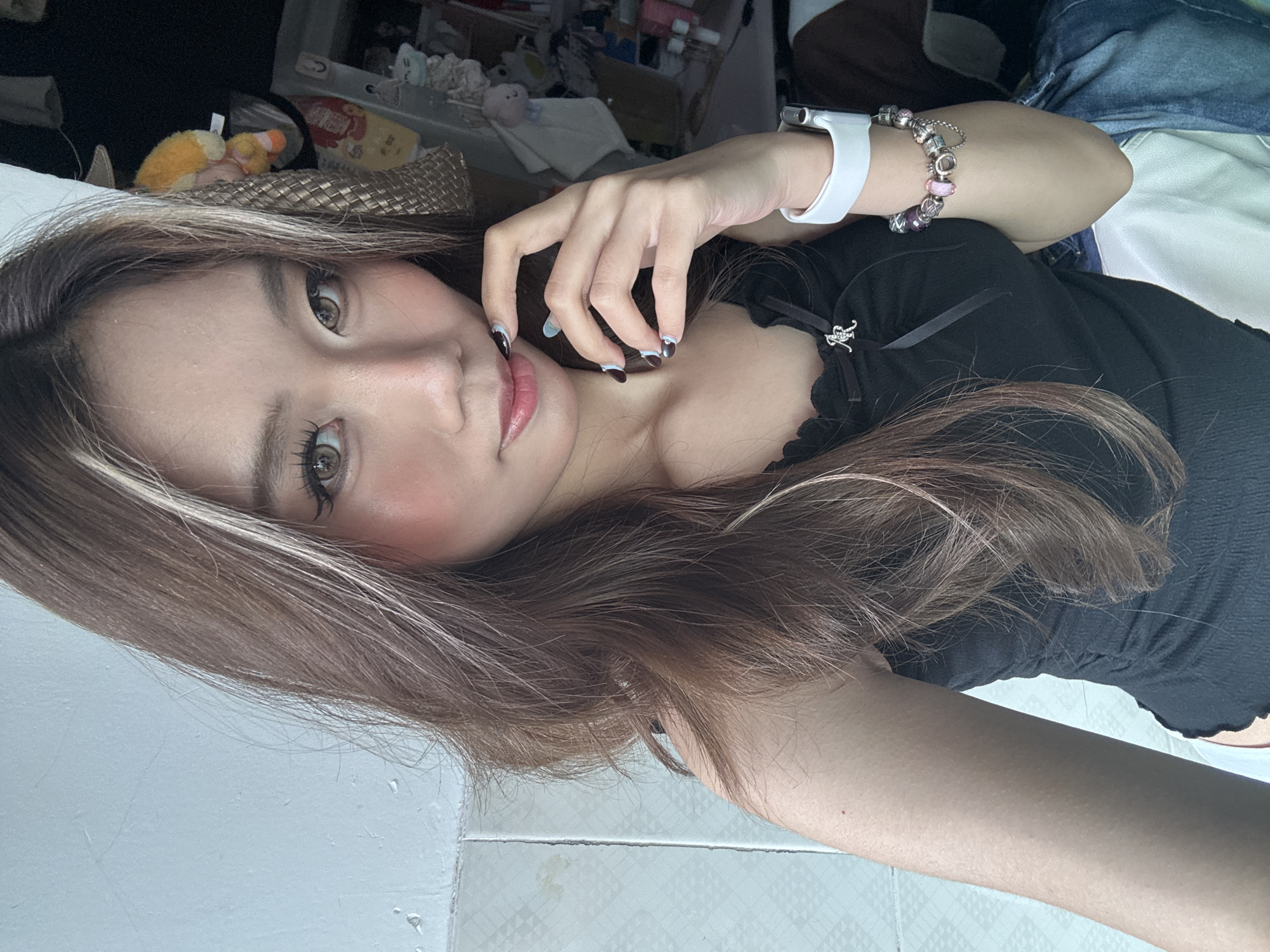 A selfie image, showing a young woman with long brown hair and fair skin. She is wearing a black top. A white smartwatch is on her left wrist, along with a bracelet. Her fingernails are painted dark with a light-colored accent near the cuticle. The background includes a cluttered area with various objects, including stuffed toys and containers. The flooring is white and tiled. There is no discernible information to determine the exact location in Nan'an District, China.