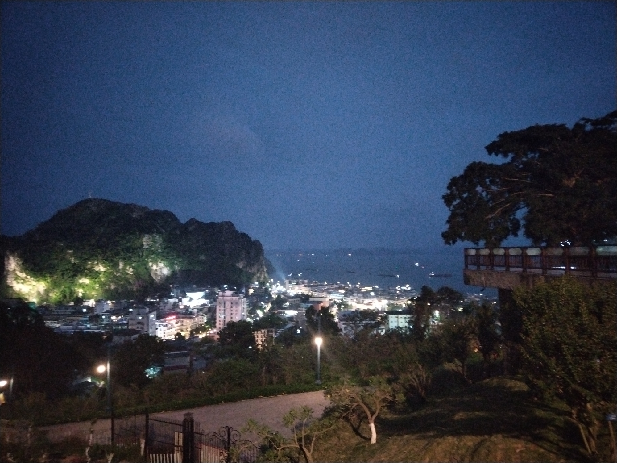 The image captures a night cityscape, likely within Hong Gai Ward, Vietnam. In the foreground, a paved path is visible, bordered by trees and shrubbery, suggesting a park or elevated viewpoint. A wrought-iron fence partially obscures the view. Above the path, the cityscape stretches into the midground, with illuminated buildings and streetlights defining the urban area. In the distance, a large, dark hill or mountain is seen on the left side, partially illuminated by lights. Beyond the city, a body of water is dimly visible, with scattered lights likely indicating boats or distant settlements. The sky is dark, with an indigo hue, suggesting either dusk or a very dark night. A structure that appears to be a raised viewing platform or pavilion with trees around it is visible to the right of the cityscape. No people are explicitly visible in the image.