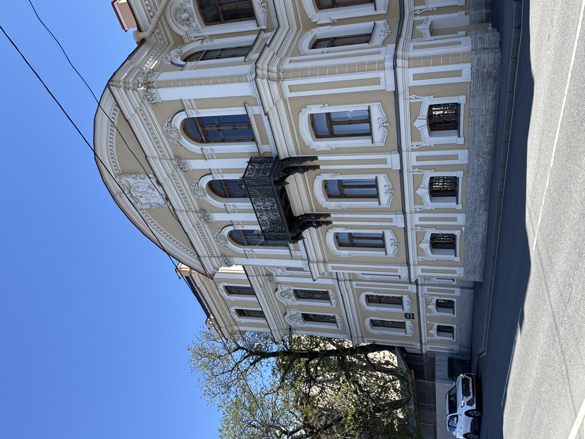 A low-angle shot captures the facade of a baroque building in Saint Petersburg, Russia, against a clear blue sky. The building is characterized by its ornate detailing, including decorative window frames, a wrought-iron balcony, and molded flourishes. A mature tree with budding leaves partially obscures the lower portion of the building. A section of a road is visible in the foreground, along with parked vehicles.