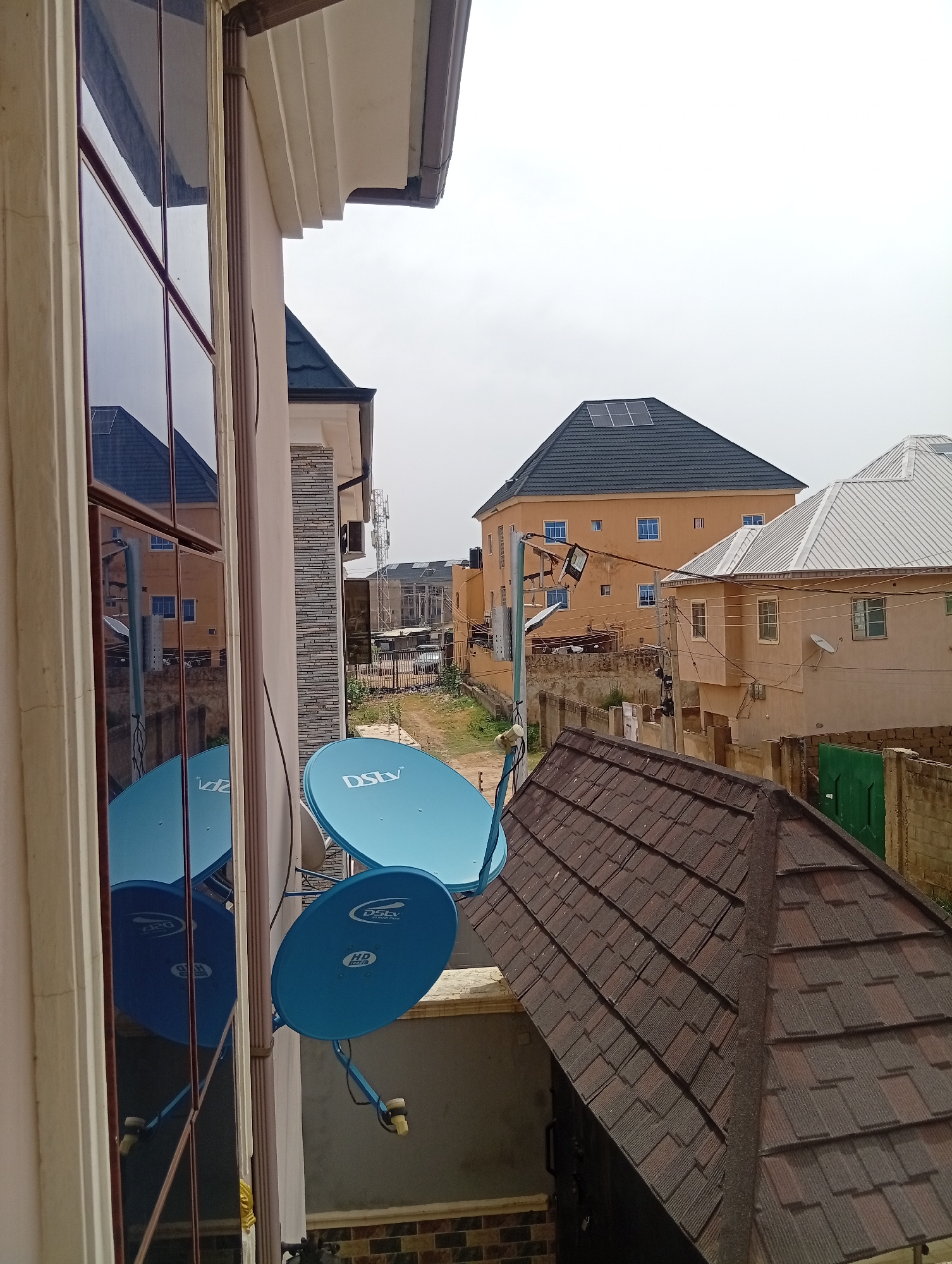The image depicts an outdoor scene in Aviation Quarters, Nigeria, likely taken from an elevated position, such as a balcony or upper floor. The foreground contains two blue DStv satellite dishes mounted on a wall. A window reflects the buildings across the street and the satellite dishes themselves. A brown tiled roof is also in the foreground.
In the background, several residential buildings are visible. One has an orange facade with blue-trimmed windows and a black roof with solar panels installed. Adjacent to it is a beige building with a silver corrugated metal roof. A green door is visible on an outbuilding near a dilapidated stone wall. A road or walkway cuts through the middle of the scene, and several utility poles and power lines run between the buildings. The overall environment appears urban, with a mix of well-maintained and older structures. The sky is overcast, suggesting daytime.