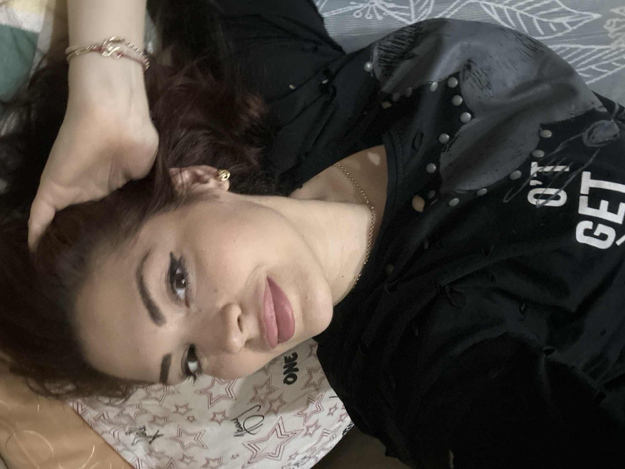A woman is lying down, looking at the camera. She has dark hair and is wearing a black top with some silver accents. She is wearing an earring. Her hand is resting on her head. The background consists of bedding with a star pattern. The location is not possible to determine from this image.