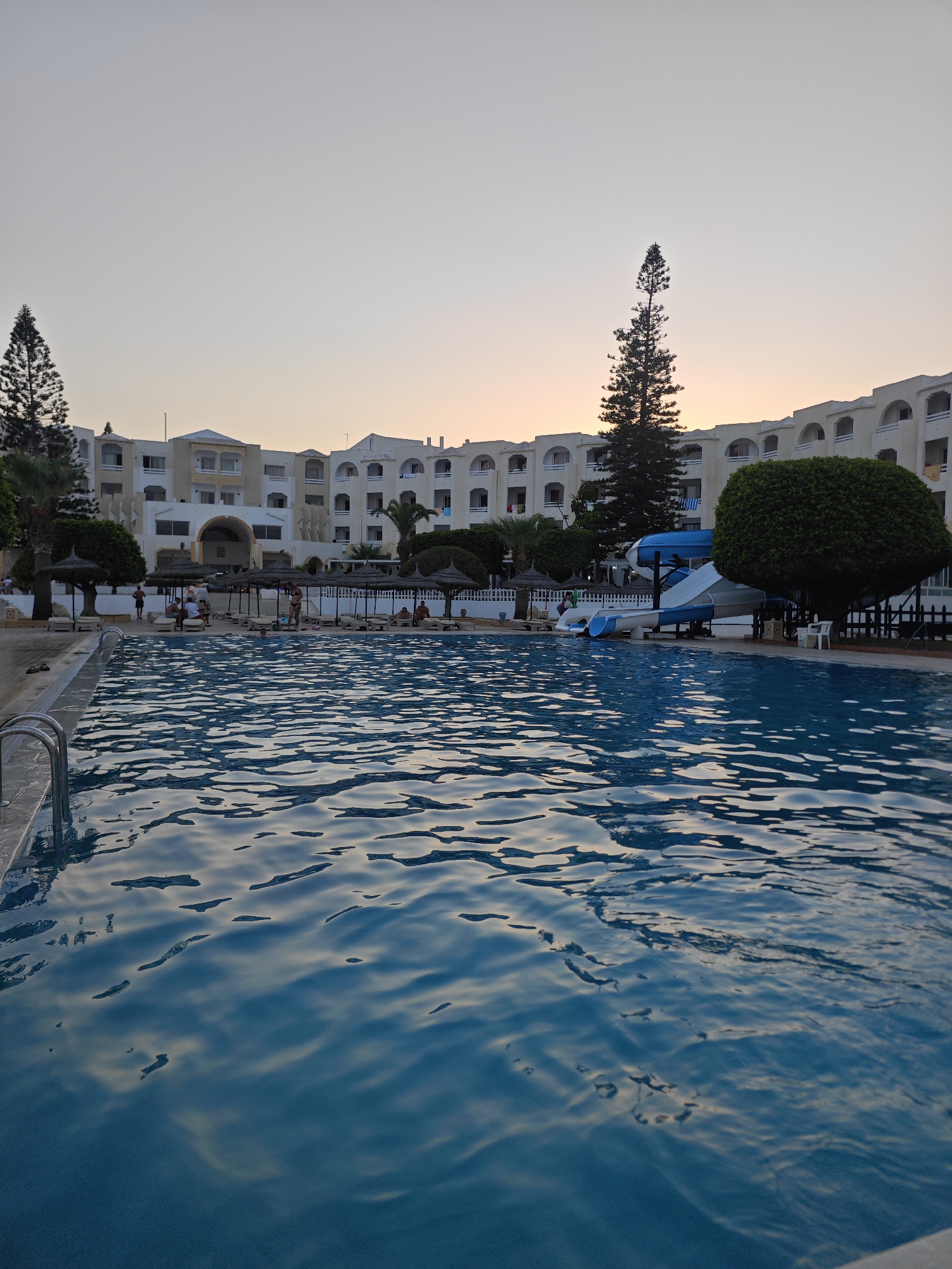 A wide-angle, low-angle shot captures a resort swimming pool at dusk in Mahdia, Tunisia. The foreground is dominated by the shimmering blue water of the pool, reflecting the soft evening sky. The far side of the pool is lined with lounge chairs, some occupied by people, and thatched umbrellas. Beyond the pool area, a multi-story white resort building with arched balconies stretches across the background, partially obscured by lush green trees. A tall, slender pine tree stands prominently to the left of the building. A blue and white water slide structure is visible to the right of the main resort building. The sky is a pale gradient of grey and soft orange, indicating the sunset. The scene depicts a peaceful resort setting, likely a hotel or vacation complex, on the coast of Mahdia.