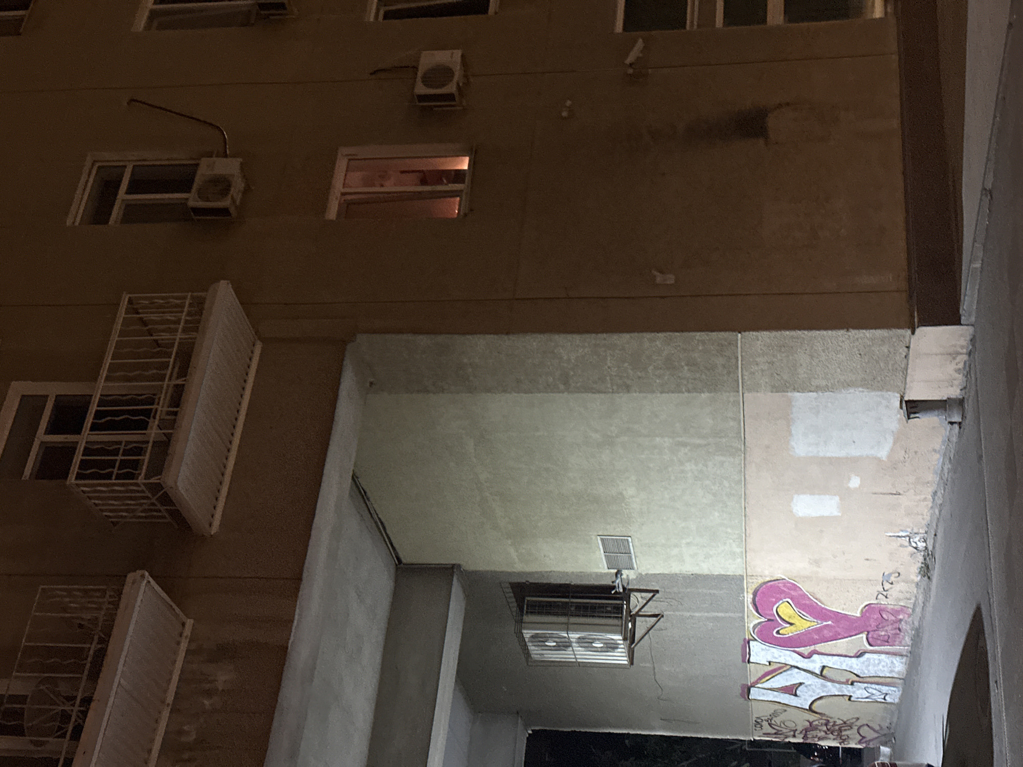 The image depicts the exterior of a multi-story building at night in Saratov, Russia. The building features balconies with metal railings on upper floors, and air conditioning units are mounted on the exterior walls. Some windows are illuminated from within, casting a warm glow. The lower portion of the building's facade is adorned with graffiti, including stylized lettering and a heart shape. A sidewalk runs along the base of the building, and shadows from overhead structures are visible on the ground. The scene is dimly lit, suggesting it is nighttime or a very overcast day.
