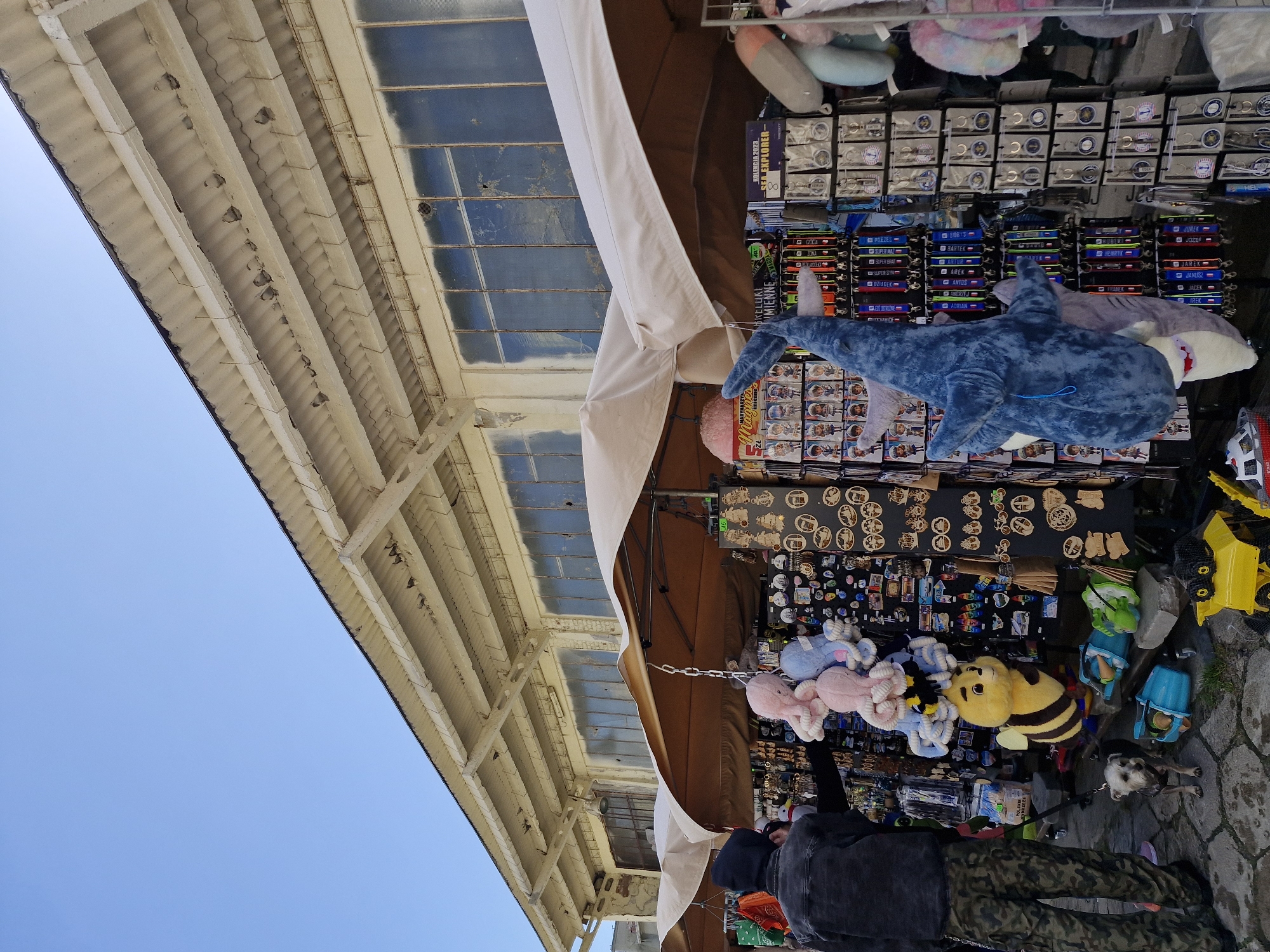 A person wearing a black hoodie and camouflage pants stands at a street market stall in Hel, Poland. They are looking at merchandise, which includes stuffed animals, keychains, and other souvenirs. A small dog stands at their feet. The stall is set up under a tent, and behind it is a weathered building with large windows. The sky is clear and blue.