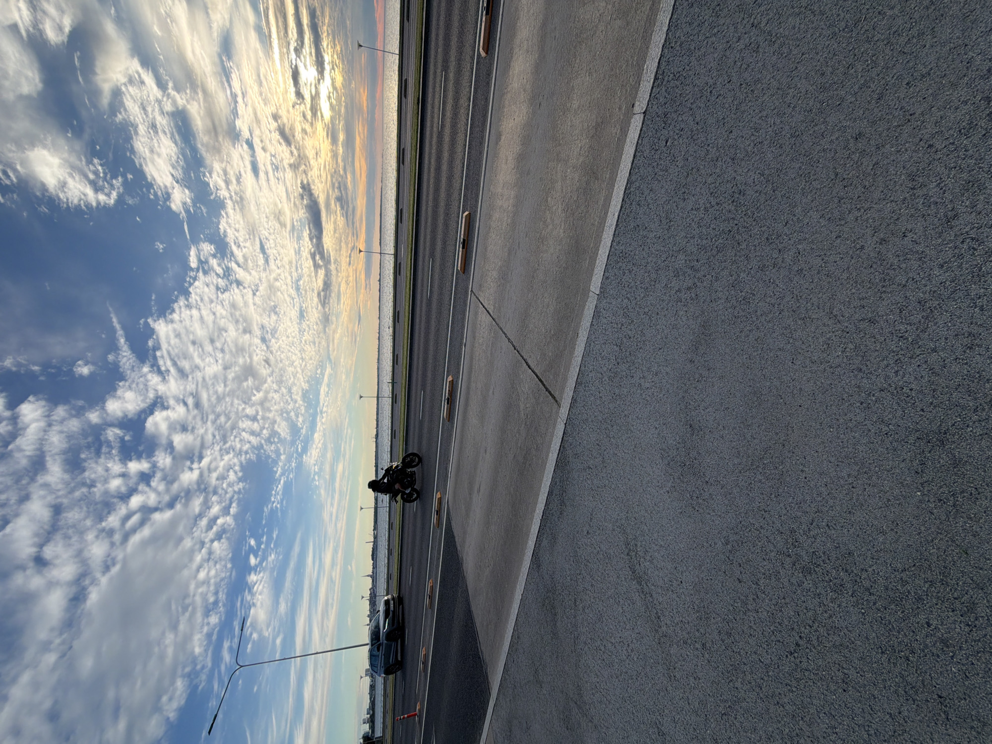 A motorcycle rider is silhouetted against a cloudy sky on a road adjacent to a body of water. A car is also visible on the road. The scene appears to be a coastal area in Tallinn, Estonia, possibly near the Pirita Road or a similar thoroughfare. The sky is a mix of blue and white clouds, with hints of sunset colors on the horizon. The road is paved, with markings indicating lanes and dividers. There are also streetlights and what appear to be speed bumps or traffic calming devices along the road. The overall atmosphere is calm and serene, with a sense of movement from the vehicles on the road.