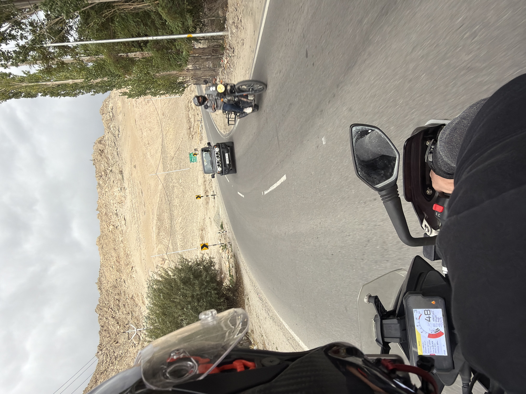 The image, taken from the perspective of a motorcycle rider, depicts a road in or near Spituk, India. The road curves to the left, with a barren, rocky mountain range in the background. Tall trees line the right side of the road. Ahead on the road, a black SUV is driving, followed by a motorcycle with a rider wearing a helmet. The motorcycle rider's helmet visor is visible in the foreground. The motorcycle's digital dashboard displays a speed of 48 and other information. The sky is overcast.