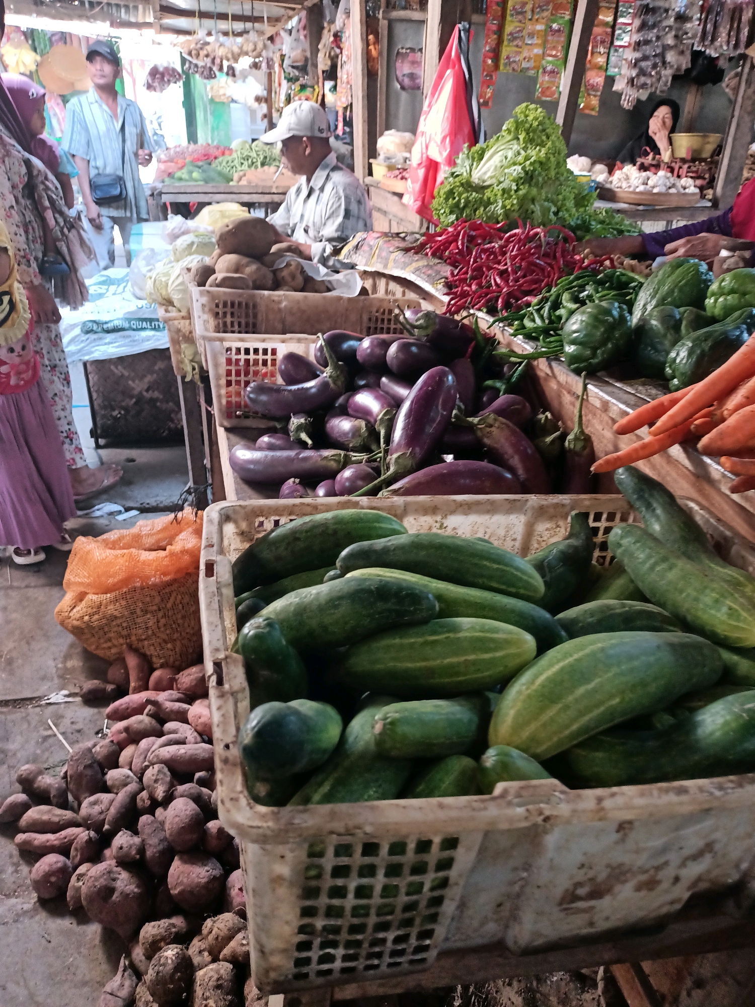 This is a photograph of a busy marketplace in Badas, Indonesia. In the foreground, several baskets overflowing with fresh vegetables are displayed. One basket contains numerous green cucumbers, some large and others smaller. To the left of the cucumbers, a large orange mesh bag holds sweet potatoes, with more scattered on the ground beneath. Further back, a basket is filled with purple eggplants. Beyond the vegetables, a man wearing a white and blue striped shirt and a black cap is standing, holding a black bag. He appears to be interacting with a woman in a colorful dress and a headscarf to his left. Behind them, more market stalls are visible, with various produce and goods displayed. Another man, wearing a white shirt and a white cap, is seated and smoking a cigarette. In the background, shelves are stocked with colorful items, possibly spices or packaged goods, and hanging decorations are visible. The overall scene depicts a vibrant, open-air market with a variety of fresh produce and people engaged in commerce.