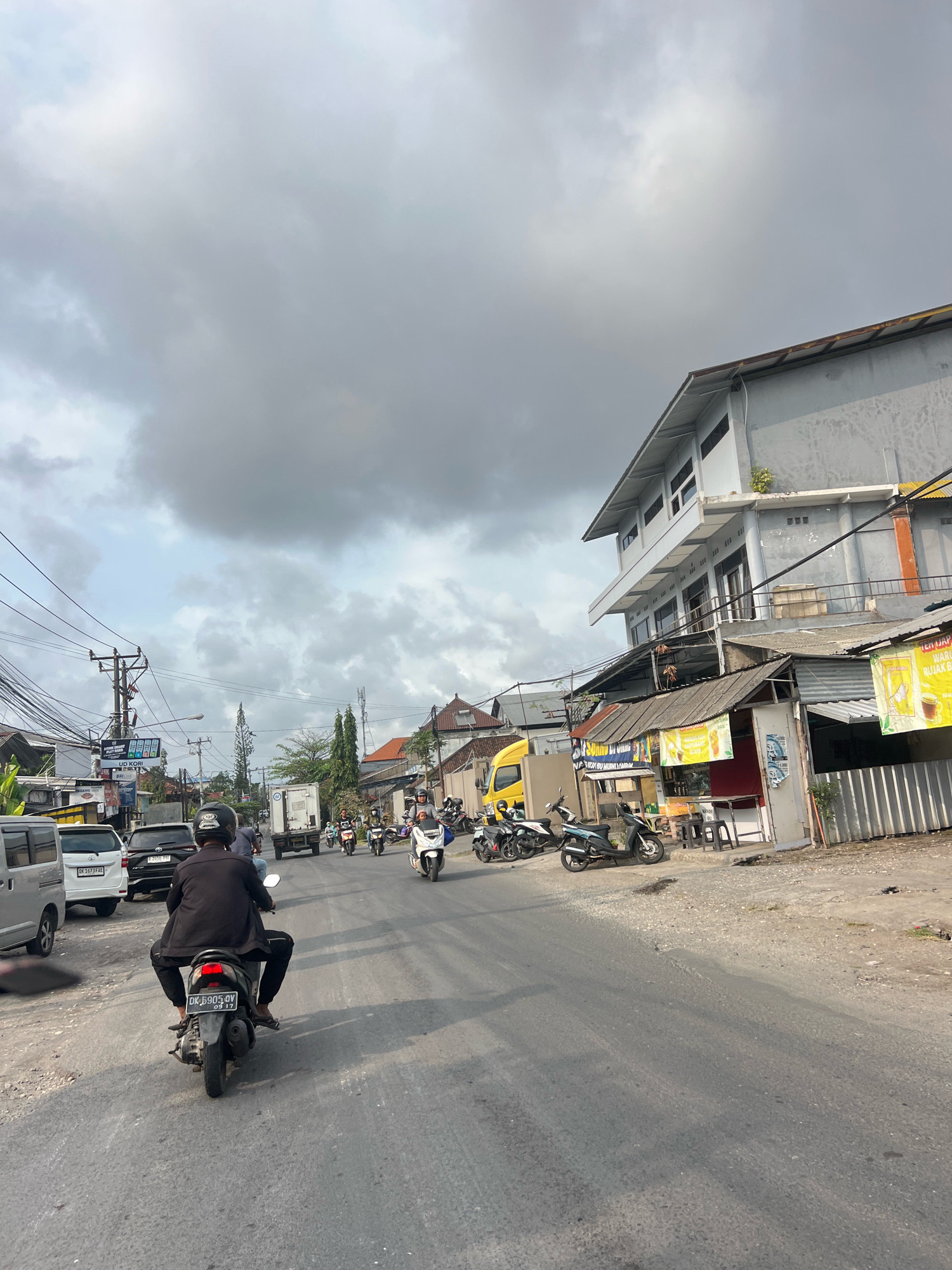 This is a street-level, eye-level, vertical, medium shot captured on an overcast day in Denpasar, Indonesia. The scene depicts a moderately busy street with various vehicles and buildings. In the foreground, a person is riding a scooter from behind, wearing a black jacket and black pants. The scooter has a license plate that reads "DK 6905 QY" and "09 17". Other vehicles, including cars and multiple scooters, are present on the road, moving in both directions. On the right side of the street, there are buildings, including a two-story structure with a gray facade and several smaller shopfronts with awnings and advertisements. The sky is heavily clouded with gray tones, suggesting an impending or recent rain. Power lines and utility poles are visible along the street, contributing to the urban environment. The road surface appears to be asphalt with some patches of dirt and debris along the sides.