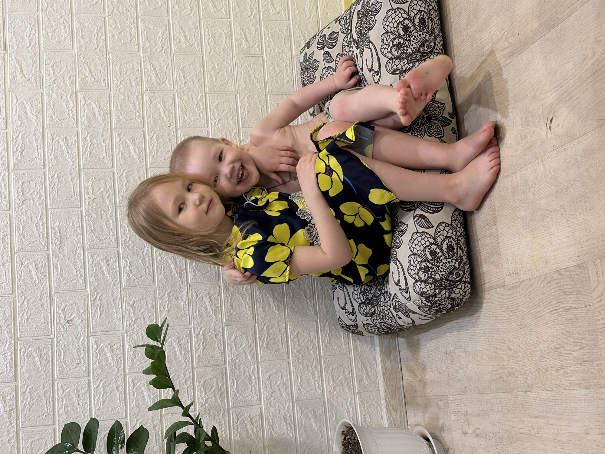 Two young children are lying on a patterned cushion on a light-colored wooden floor. The child on top is fair-skinned, possibly a boy, with a small amount of hair. He is shirtless and smiling. The child below appears to be a girl with light brown, shoulder-length hair. She is wearing a dark-colored dress with yellow floral print and is looking at the camera. The background features a textured white wall. A houseplant in a white pot is in the lower left corner of the image. The location is Noviy, Russia.
