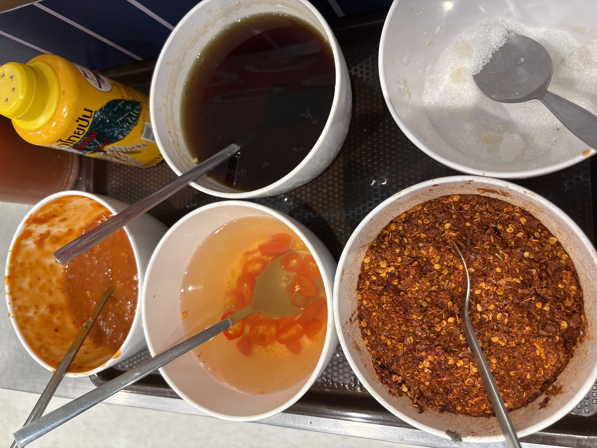 The image shows a selection of Thai condiments in white bowls arranged on a tray. There's a yellow bottle of Thai condiment (likely soy sauce or a similar seasoning). The bowls contain a dark sauce (likely soy sauce or a fish sauce based sauce), granulated white sugar, a red chili sauce, chilies in vinegar, and crushed dried chilies. Silver spoons are placed inside each bowl. The location appears to be a restaurant in บ้านบางรักษ์, Thailand, as the Thai condiments are commonly used in Thai cuisine.