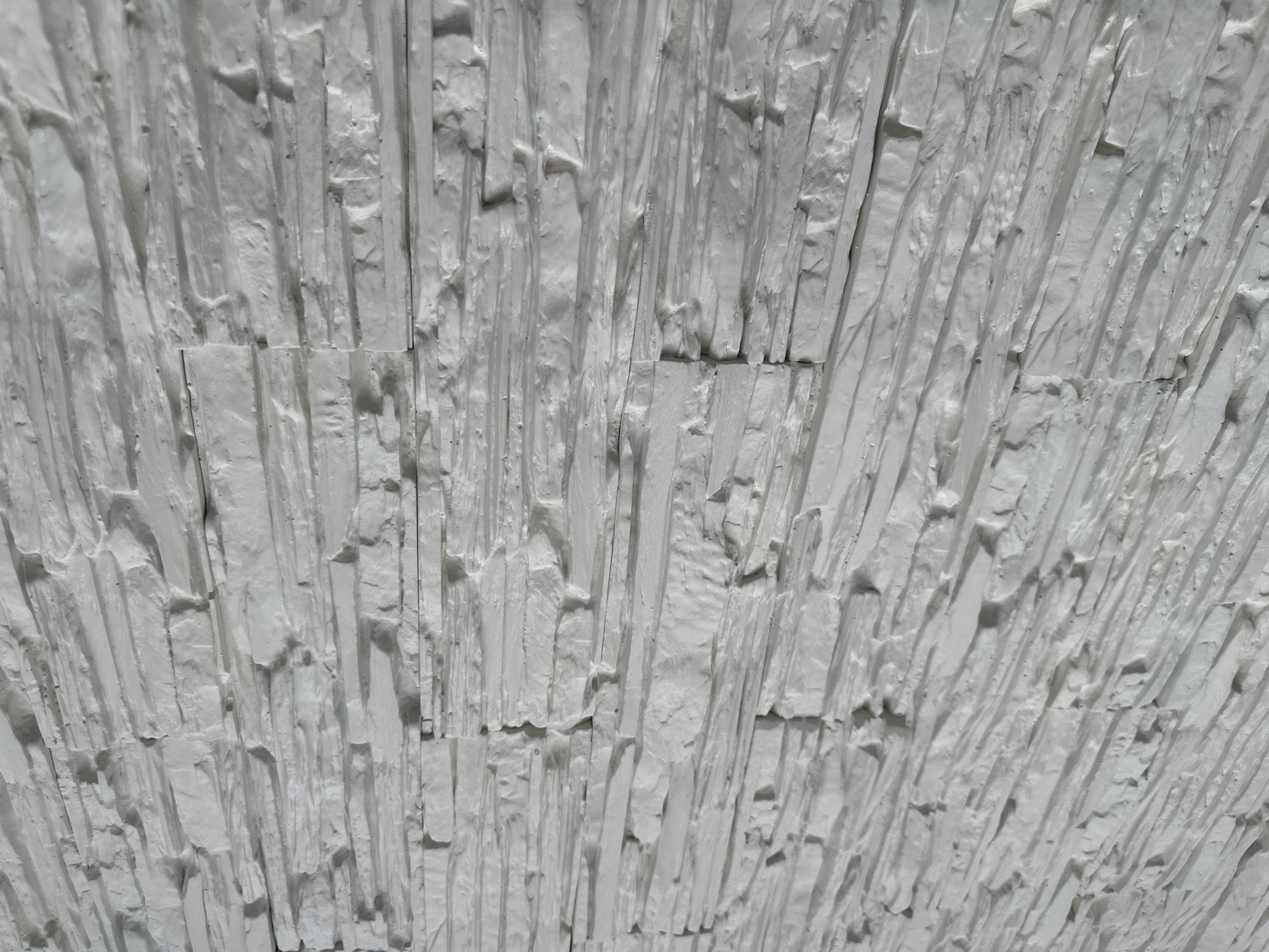 The image shows a close-up of a white, textured surface. It appears to be a wall or architectural feature composed of roughly rectangular pieces arranged vertically. The texture is uneven and rugged, with visible ridges, grooves, and variations in the surface of each piece. There is no visible indication of location in the city of Unknown, Unknown. No people, subjects, objects, actions, or interactions are discernable other than the texture on a white wall.