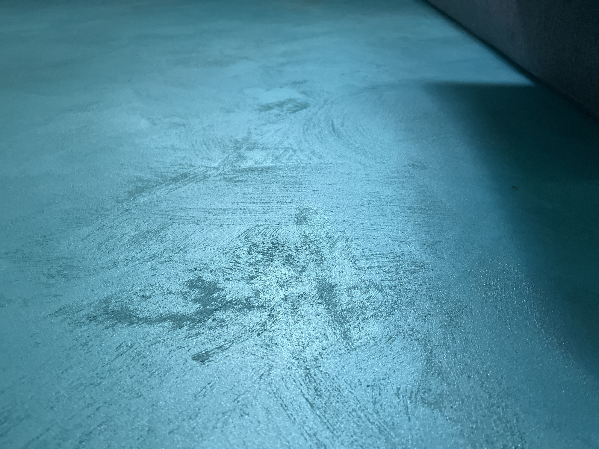 The image displays a close-up view of a textured surface, predominantly in a teal or turquoise color, seen from a low angle looking upwards and to the right. The surface exhibits a distinct, irregular texture with visible brush-like strokes and swirling patterns, suggesting it could be painted concrete, a stucco wall, or a heavily textured fabric. Variations in light and shadow highlight the unevenness of the surface, creating lighter, reflective areas and darker, recessed patches.
In the upper right portion of the frame, a dark, indistinct object is partially visible, appearing as a rolled edge or corner of something, possibly a mat or fabric. This object casts a soft shadow onto the teal surface directly beneath it. No people or specific activities are depicted. The lighting is subtle, emphasizing the material's texture rather than being overtly bright. There is no visible text. The overall impression is an abstract study of color and texture.