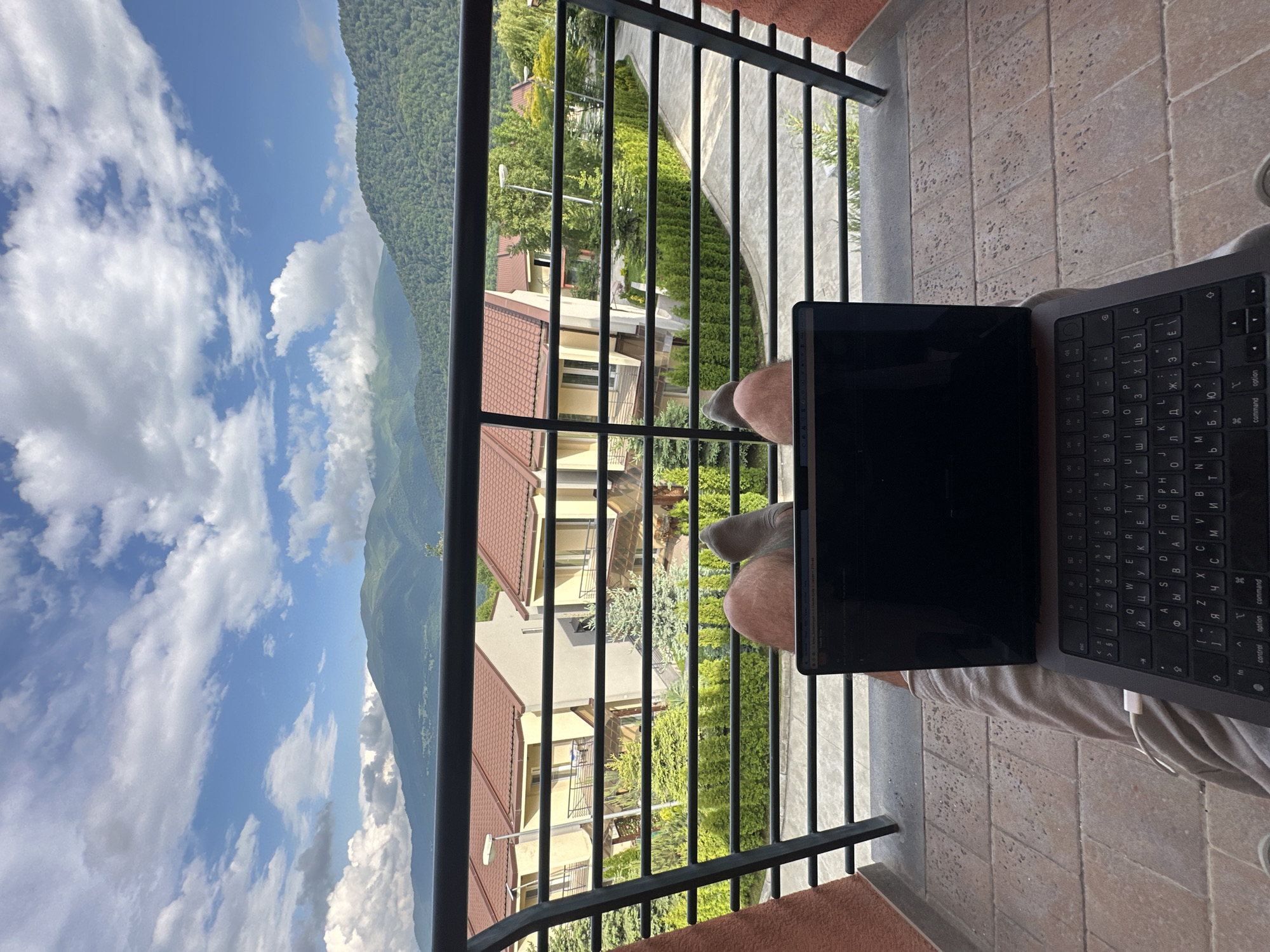 This image shows a person sitting on a balcony with a laptop in front of them. The scene is set in Dilijan, Armenia, with a view of lush green mountains and a bright blue sky with fluffy white clouds. Several houses with red tile roofs are visible below the balcony, surrounded by trees and manicured gardens. The balcony itself has a black metal railing and a tiled floor. The person is wearing light-colored shorts, and their bare legs and feet are visible. A MacBook laptop is open on the balcony, and the keyboard is visible, displaying Cyrillic characters. The time of day appears to be daytime, with bright sunlight casting shadows. The overall mood suggests a relaxed and productive atmosphere, possibly a remote work or study session in a scenic location. The visible text is primarily from the laptop's keyboard layout.