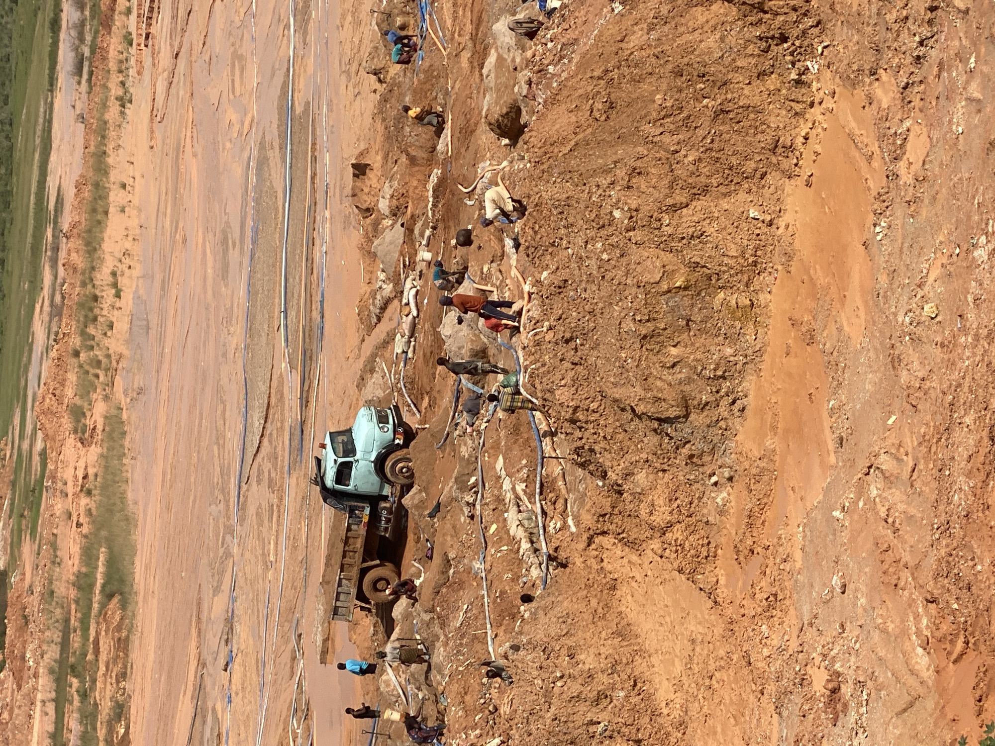 The image depicts an aerial view of a mining operation in Jos, Nigeria. The landscape is dominated by a vast expanse of reddish-brown soil, indicating a mining site. Several individuals are actively engaged in work, some near a light blue dump truck laden with soil. Others are clustered around excavated areas, appearing to be involved in manual labor, possibly digging or sifting through the earth. Blue and white pipes are visible, suggesting a water pumping or drainage system in place to manage the wet conditions of the mine. The terrain is uneven and heavily disturbed, characteristic of a site undergoing resource extraction. The overall scene conveys a sense of industrious activity within a rugged, earth-toned environment.