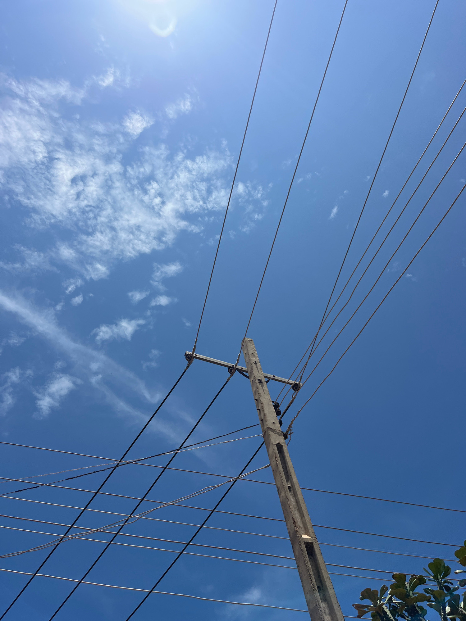 The image depicts a clear blue sky with wispy white clouds. A concrete utility pole stands prominently, angled slightly to the right, supporting a complex network of overhead wires. The wires stretch diagonally across the frame, creating a pattern against the sky. In the lower right corner, a portion of green foliage from a tree is visible, suggesting an outdoor environment, potentially within a residential or semi-urban area of Jos, Nigeria, given the presence of utility infrastructure. There are no people or visible activity in the scene.