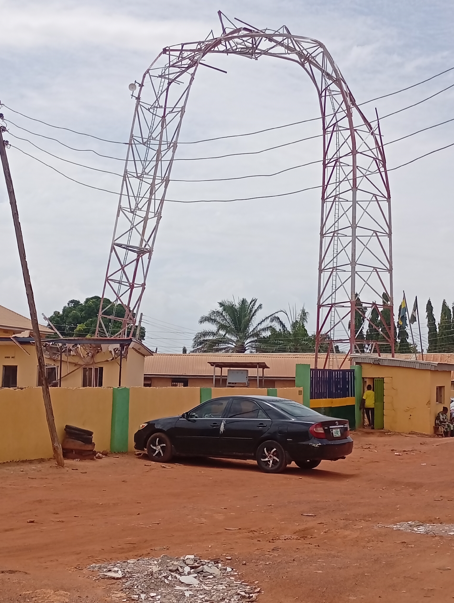 The image depicts an outdoor scene dominated by a large metal tower, possibly a communication or transmission tower, which has collapsed or bent into a dramatic arch shape. The central, sagged portion of the tower rests upon or very close to the roof of a single-story building with light yellow walls and a brown roof, causing significant damage to the roof structure. Faint text, "GENDER UNIT", is visible on the damaged building above a window.
In the foreground, a black sedan car with distinct star-shaped wheel rims is parked on a reddish-brown dirt ground, facing right. To the left, a wooden utility pole with multiple power lines runs vertically. Near the utility pole and a low yellow and green wall, several stacked tires and some wooden debris are visible. Scattered rubble lies on the dirt ground in the immediate foreground.
Behind the wall, various buildings with similar architectural styles are visible. On the right side of the image, a person wearing a yellow shirt stands in an open doorway of one of these buildings, while two other individuals are seated further to the right. The background features palm trees, other green vegetation, and several flags on poles. The sky is overcast, indicating daytime. The scene conveys an event of structural failure, likely an accident involving the tower. The location context is Sabon Tasha, Nigeria.