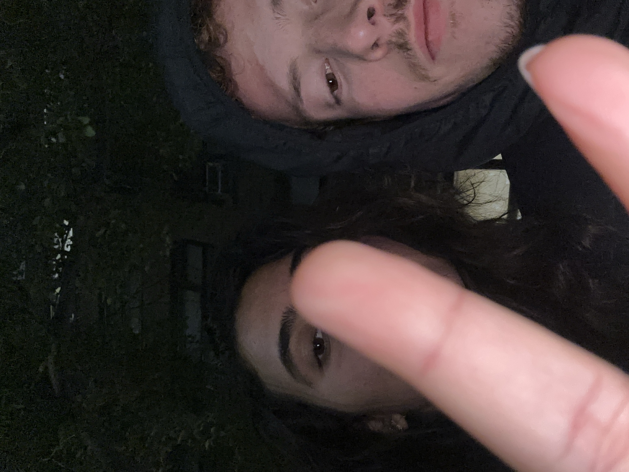 Two people are posing for a selfie. The top person is wearing a black hooded jacket. The person below is making a peace sign. The scene appears to be outdoors at night. Buildings and foliage are visible in the background. The location is the city of New York, United States of America.