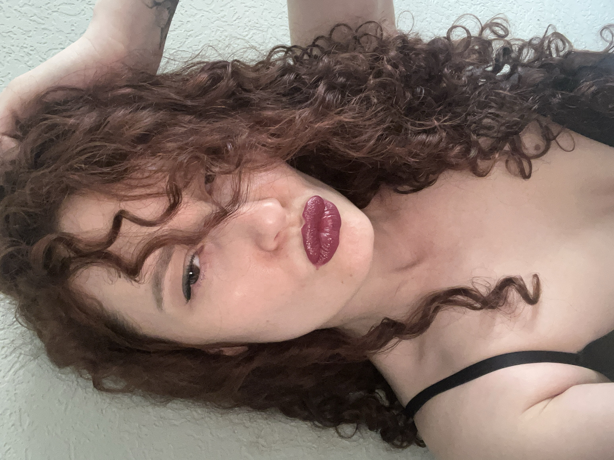 A woman with curly brown hair is lying down, facing the camera. She is wearing a dark lipstick and a black strap is visible on her shoulder. The background is a textured wall. The image does not provide information about location in Saint Petersburg, Russia.