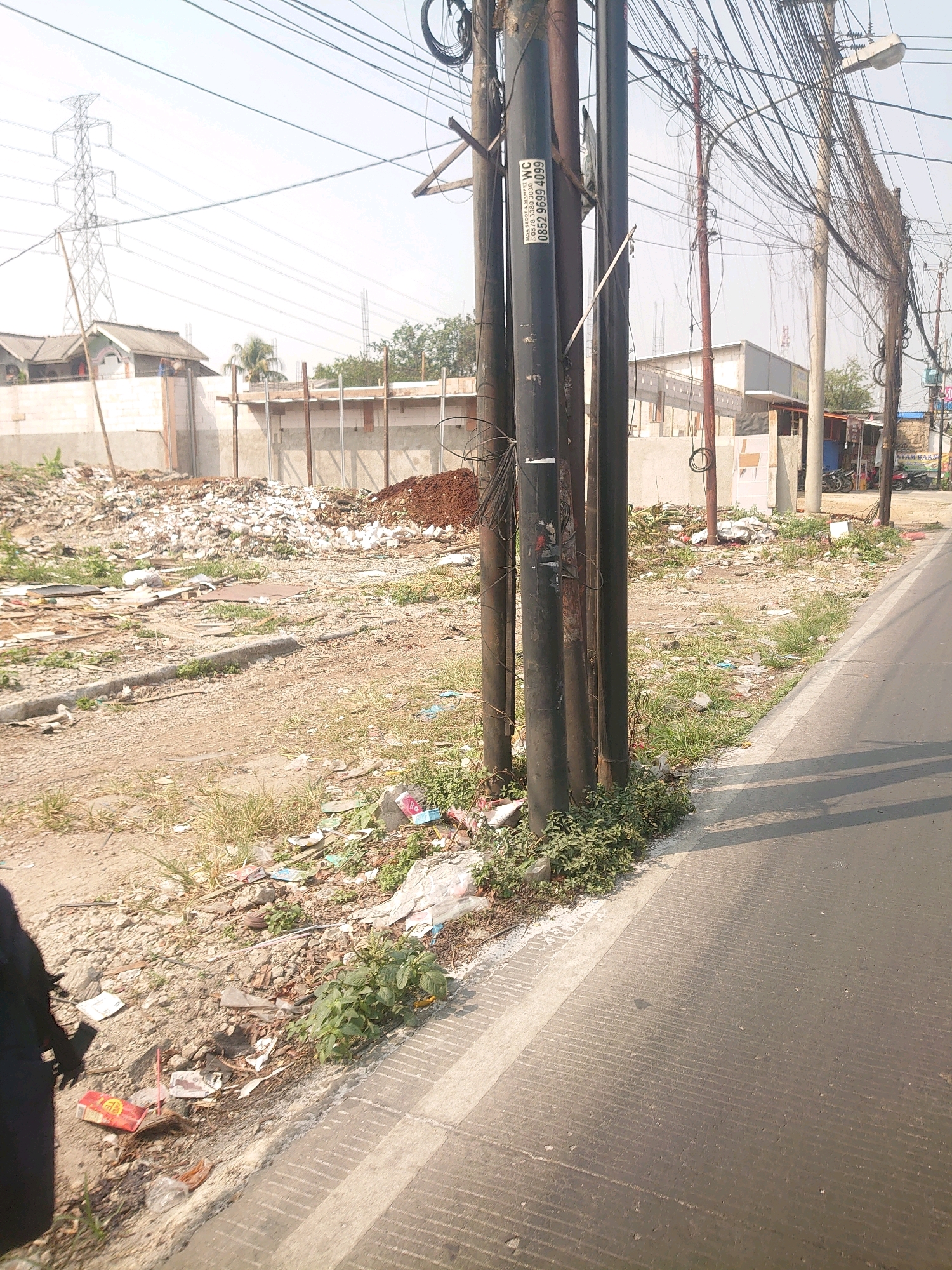 The image depicts a street scene in Bojong Gede, Indonesia, characterized by a significant amount of urban waste and debris. In the foreground, a paved road with a white line marking its edge runs from right to left. A section of unpaved ground, littered with various types of refuse including plastic bags, paper, and other discarded materials, lies adjacent to the road. Several utility poles, heavily adorned with a dense network of electrical wires, dominate the mid-ground. Beyond these poles, a construction site is visible, featuring partially built structures, piles of dirt, and more accumulated waste. In the background, large electricity pylons stand against a bright, hazy sky, and residential buildings are partially obscured by the construction and debris. No people are directly visible in the scene, and the overall impression is one of urban neglect and ongoing construction in a developing area.